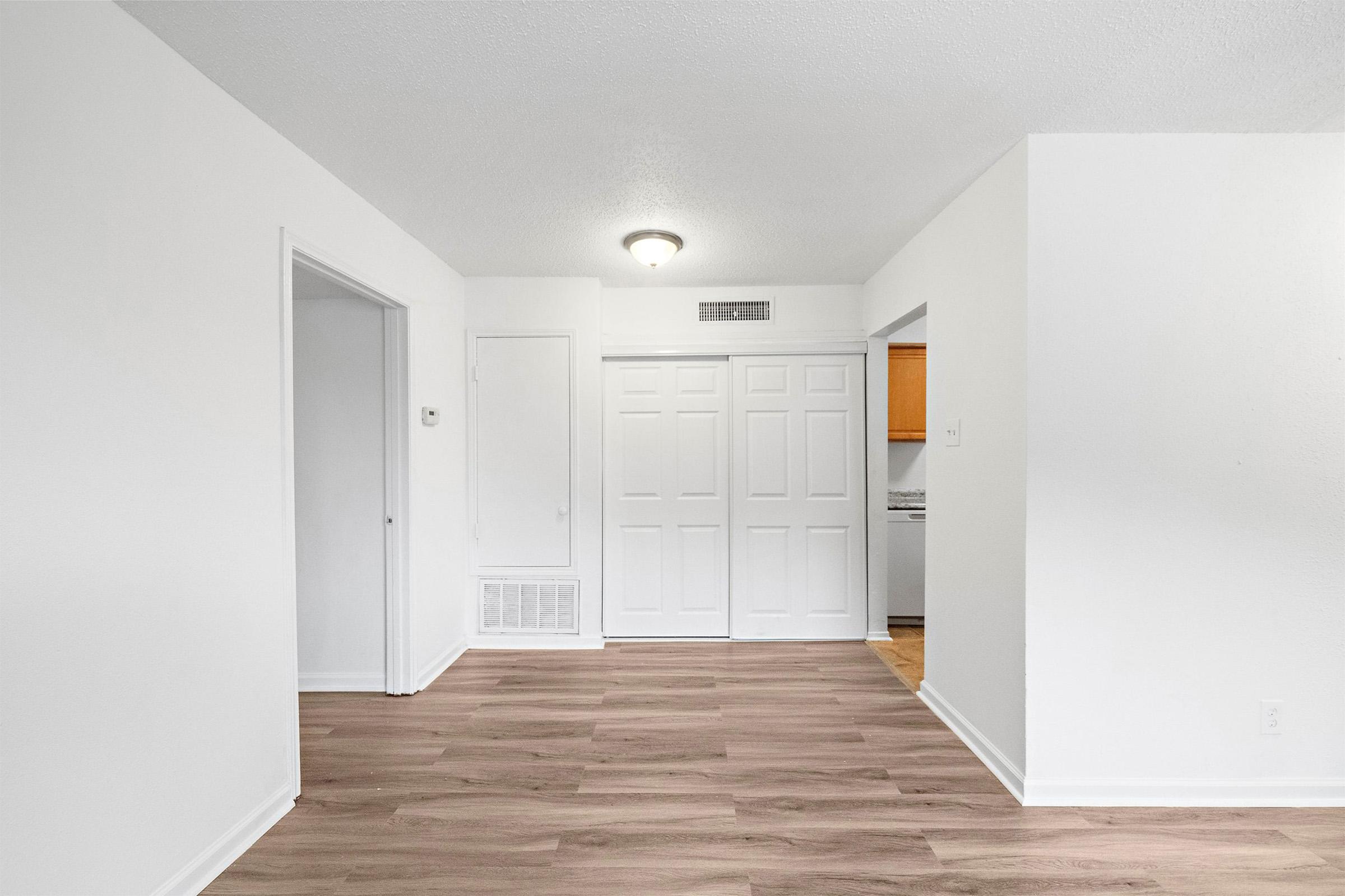A bright, empty living space featuring light-colored walls and wooden flooring. There's a closet with double doors on the left, and an open doorway leading to a kitchen area at the back. The room is well-lit with a ceiling light fixture.