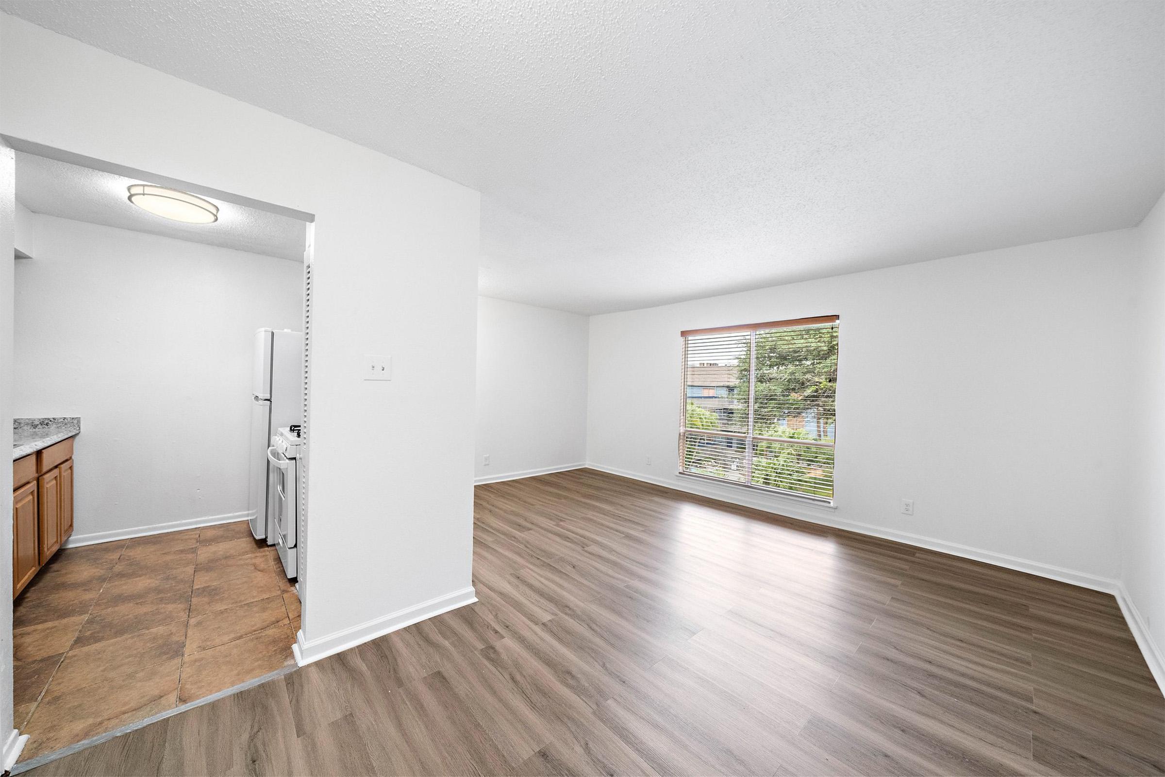 Bright and spacious room with light-colored walls and large windows providing natural light. Features a laminate floor and a doorway leading to a kitchen area with a refrigerator. The overall design is modern and airy, perfect for various interior styles.