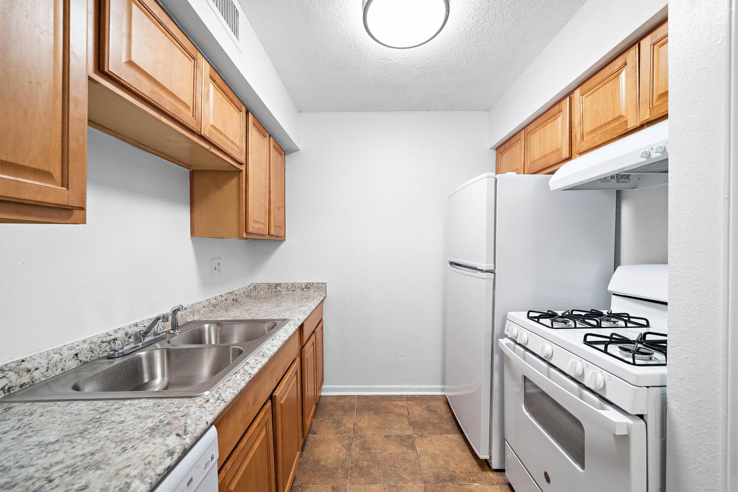 A small kitchen featuring light wood cabinets, a double sink, and granite countertops. There is a white refrigerator and a gas stove with an oven, along with a circular ceiling light. The walls are painted white, and the floor is covered with brown vinyl tiles.