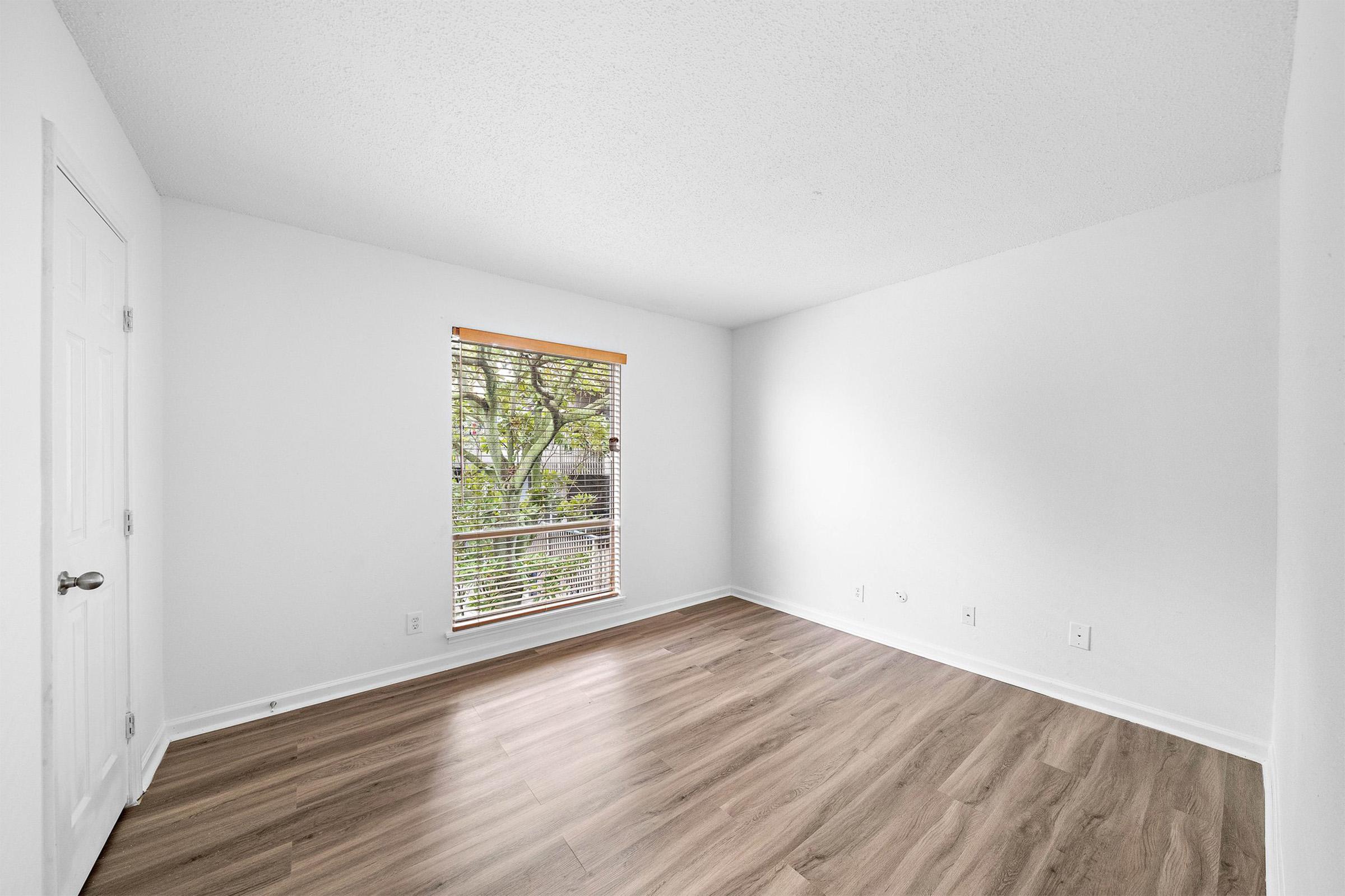 A bright, empty room with light-colored walls and a large window featuring wooden blinds. The floor is made of light wood laminate, and there are no furnishings or decorations. Outside the window, greenery can be seen, adding a natural touch to the space.