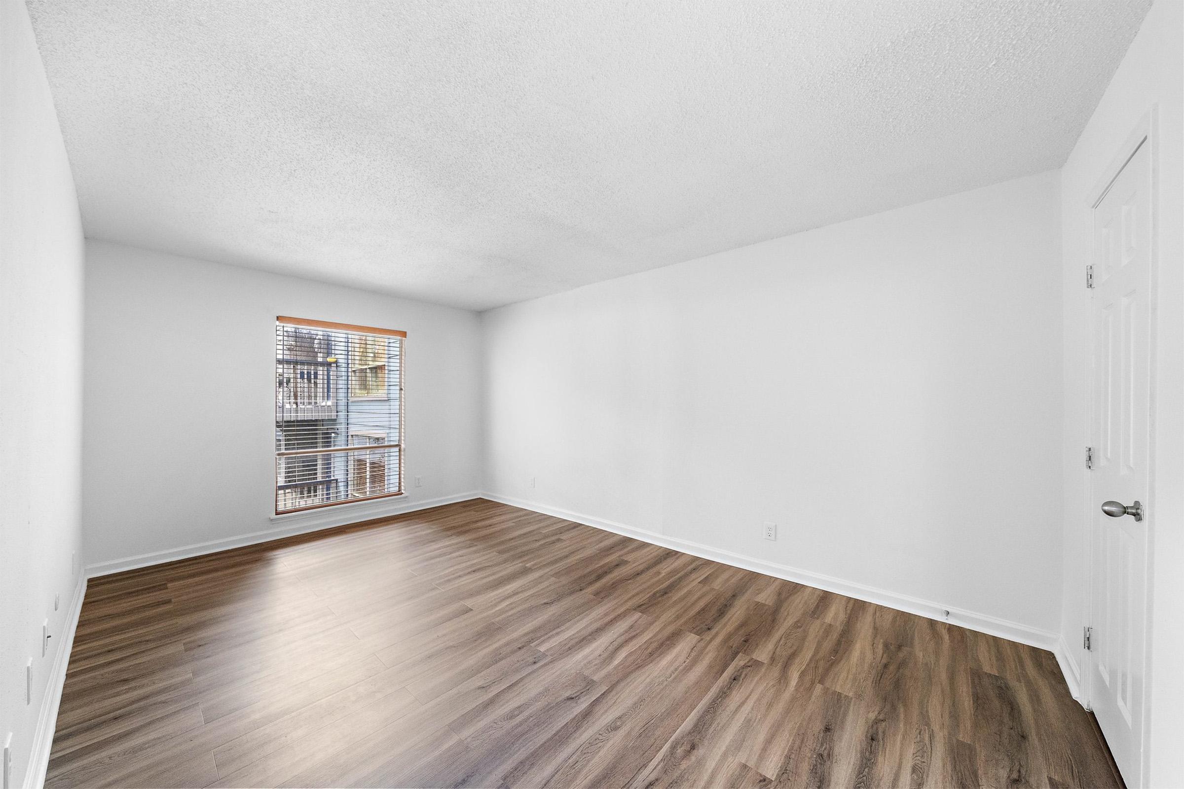 A spacious, empty room with light gray walls and a hardwood floor. A window on the left side allows natural light to enter, showcasing a view of a balcony. The room has no furniture and appears well-lit and airy, creating a bright and inviting atmosphere.