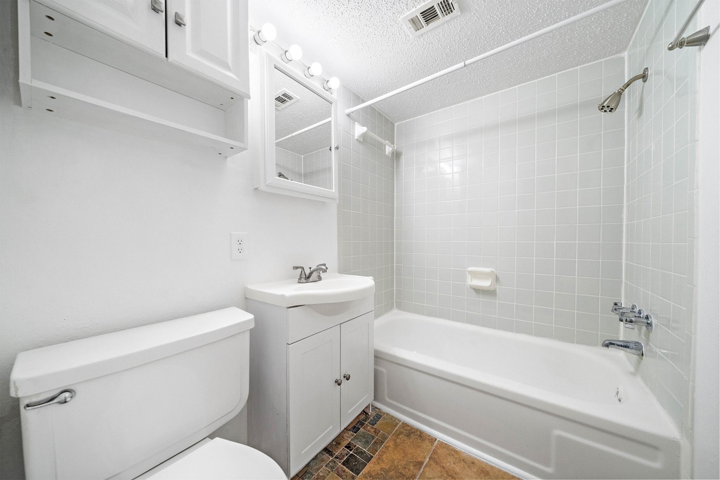 A clean and modern bathroom featuring a white bathtub, shower fixtures, a sink with a cabinet underneath, a mirror above the sink, and white cabinetry for storage. The walls are tiled in light colors, and the floor has a patterned tile design. Good lighting is provided by a fixture above the mirror.