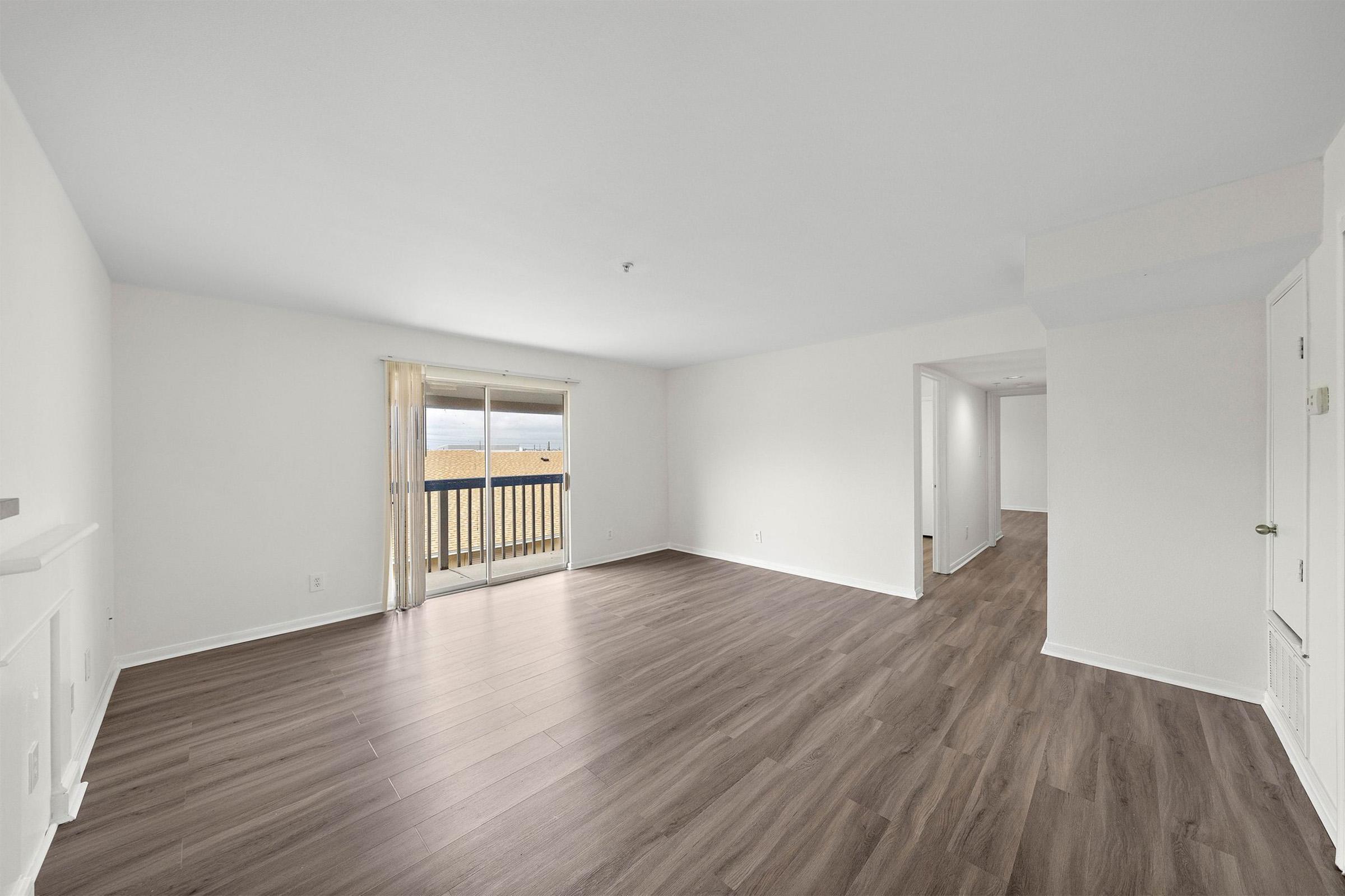 Spacious interior of an empty room featuring light-colored walls and large windows with sheer curtains. The floor is made of laminate wood, and there is a balcony with a view. The room has a modern feel with plenty of natural light, and a hallway visible leading to other areas of the space.