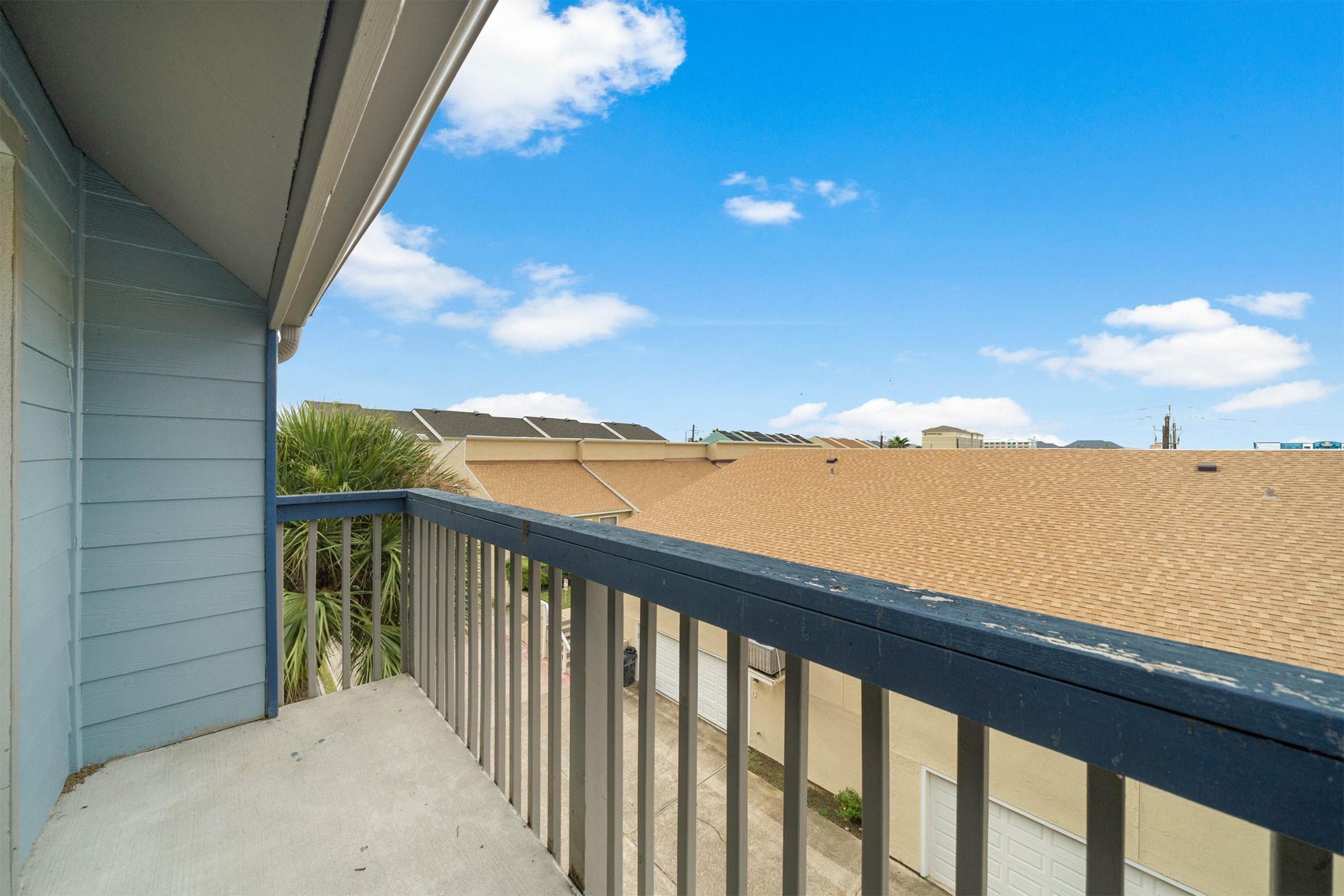 A view from a balcony overlooking rooftops, with a clear blue sky and a few clouds. In the foreground, there is a railing and concrete floor, while the background features a mix of buildings, including one with solar panels.