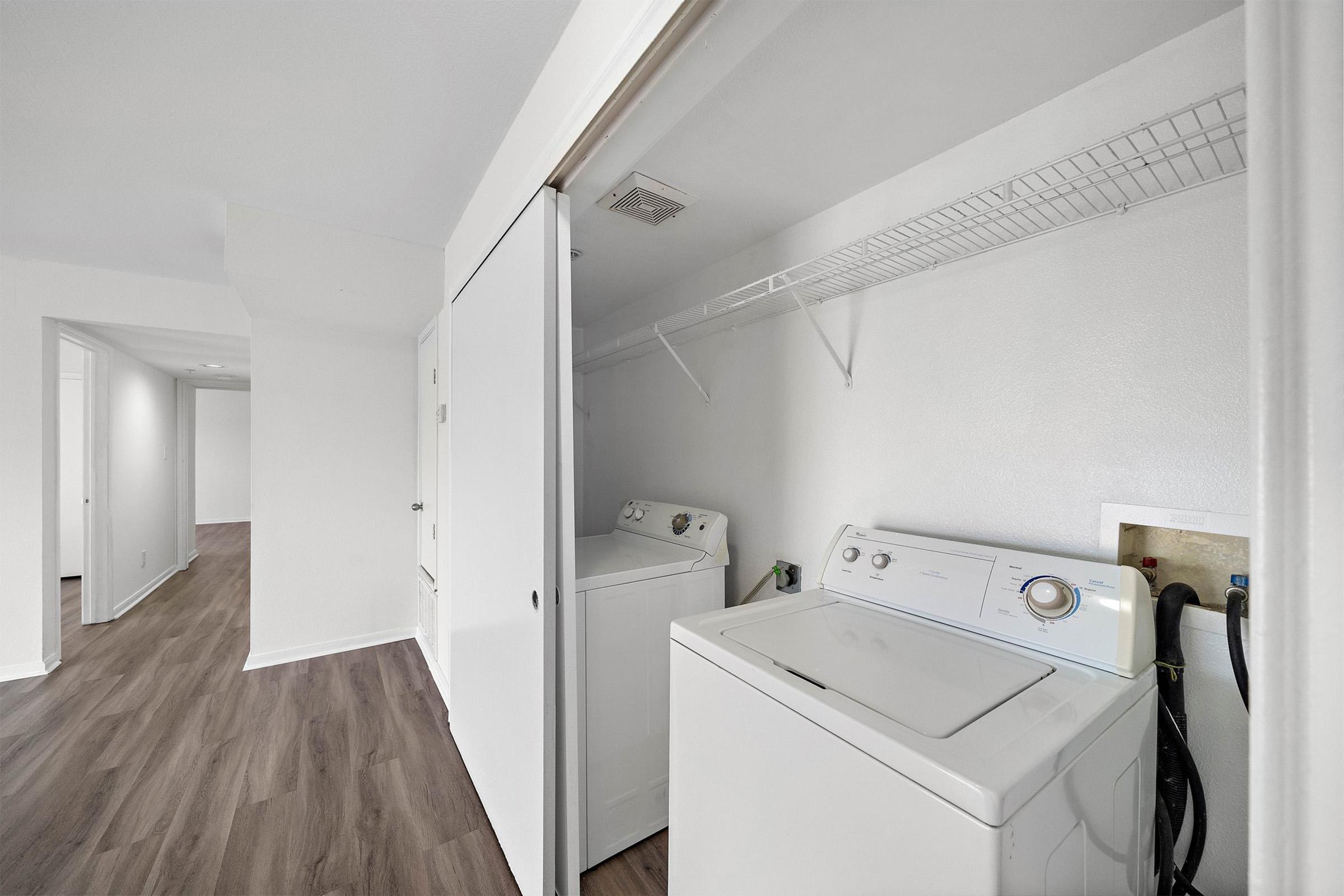 A laundry area featuring a washer and dryer next to a closet with a wire shelf. The walls are painted white, and the floor has a brown wooden appearance. A view of a hallway leading to another room is visible in the background.