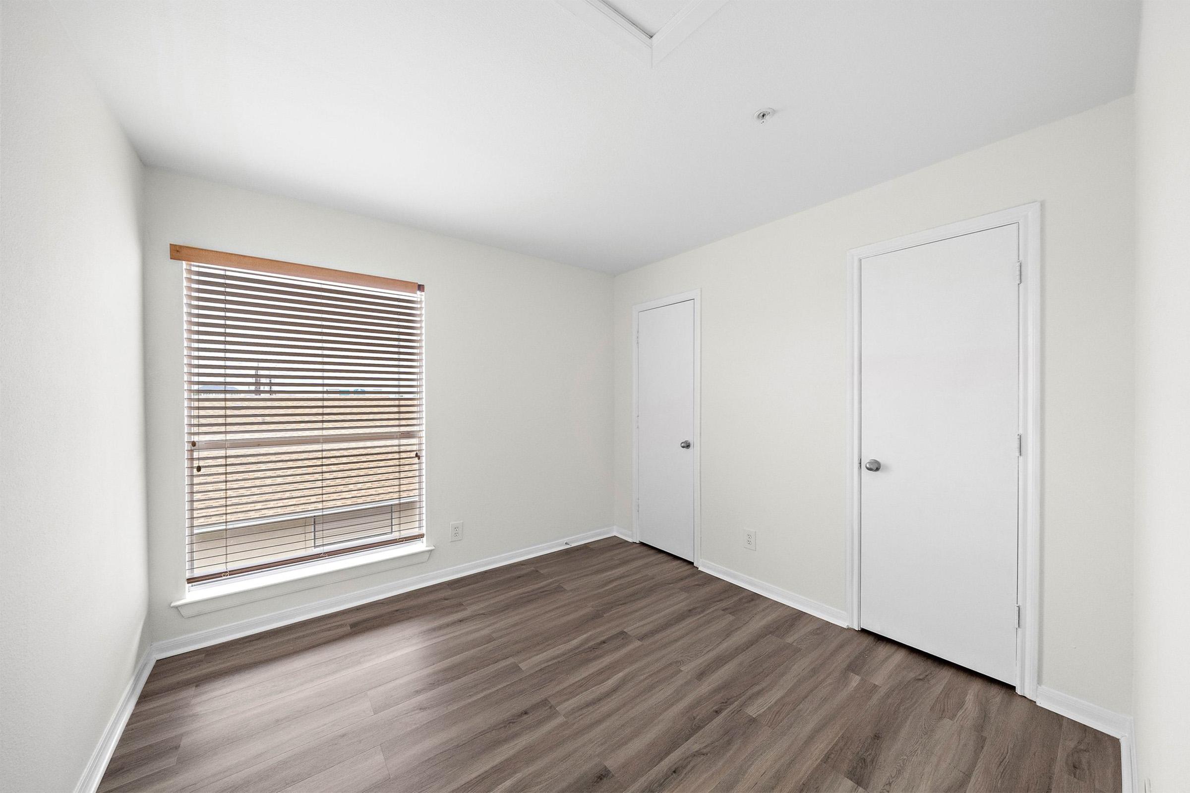 A bright, empty room featuring light-colored walls and wood-like flooring. A window with horizontal blinds allows natural light to enter, while a closed door on the right and a closet door on the left add functionality to the space. Ideal for various uses, such as a bedroom or office.