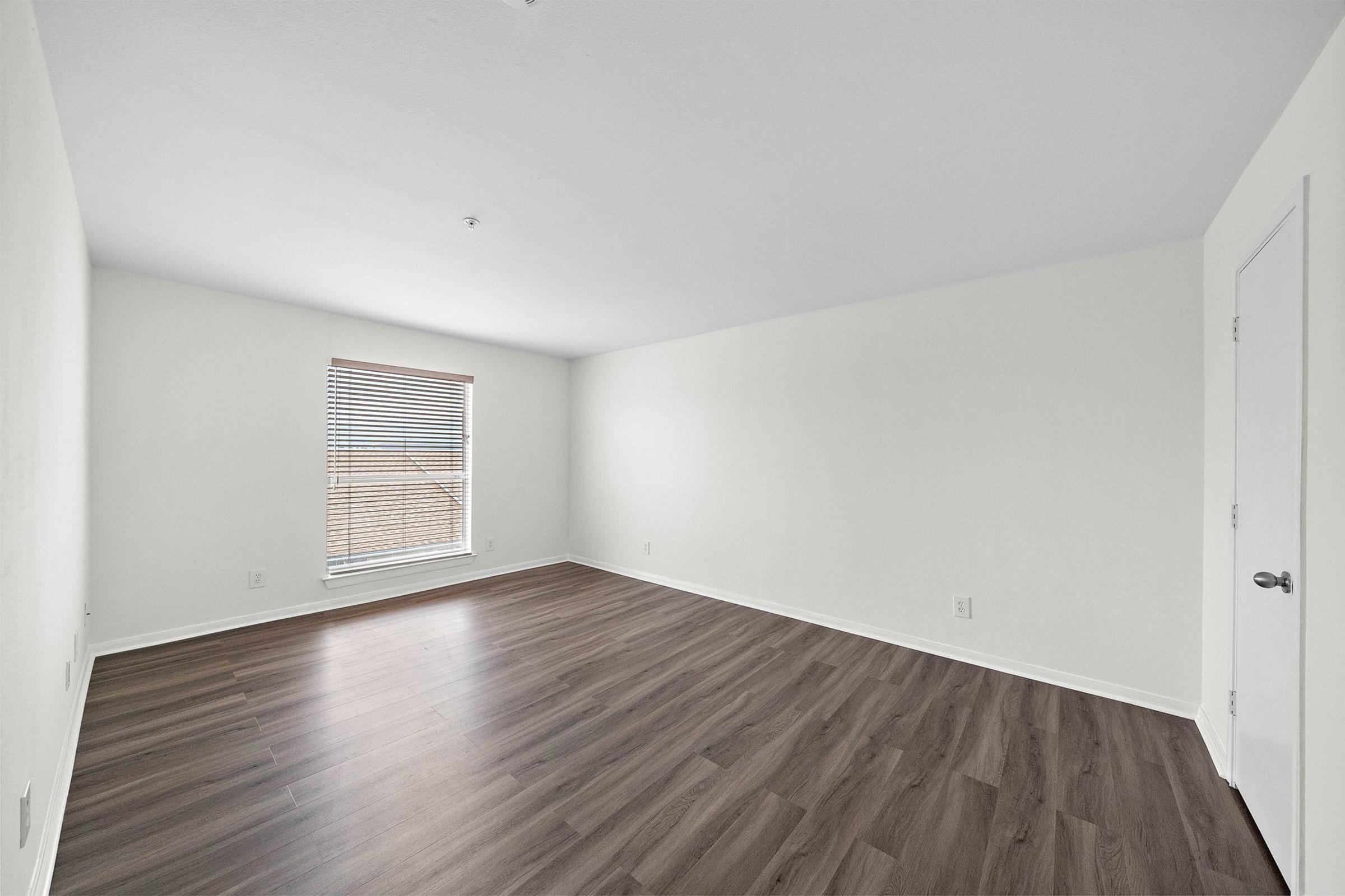 A spacious, empty room featuring light hardwood flooring, white walls, and a single window with blinds allowing natural light. The room is unadorned, offering a minimalist appearance, suitable for various arrangements or uses.
