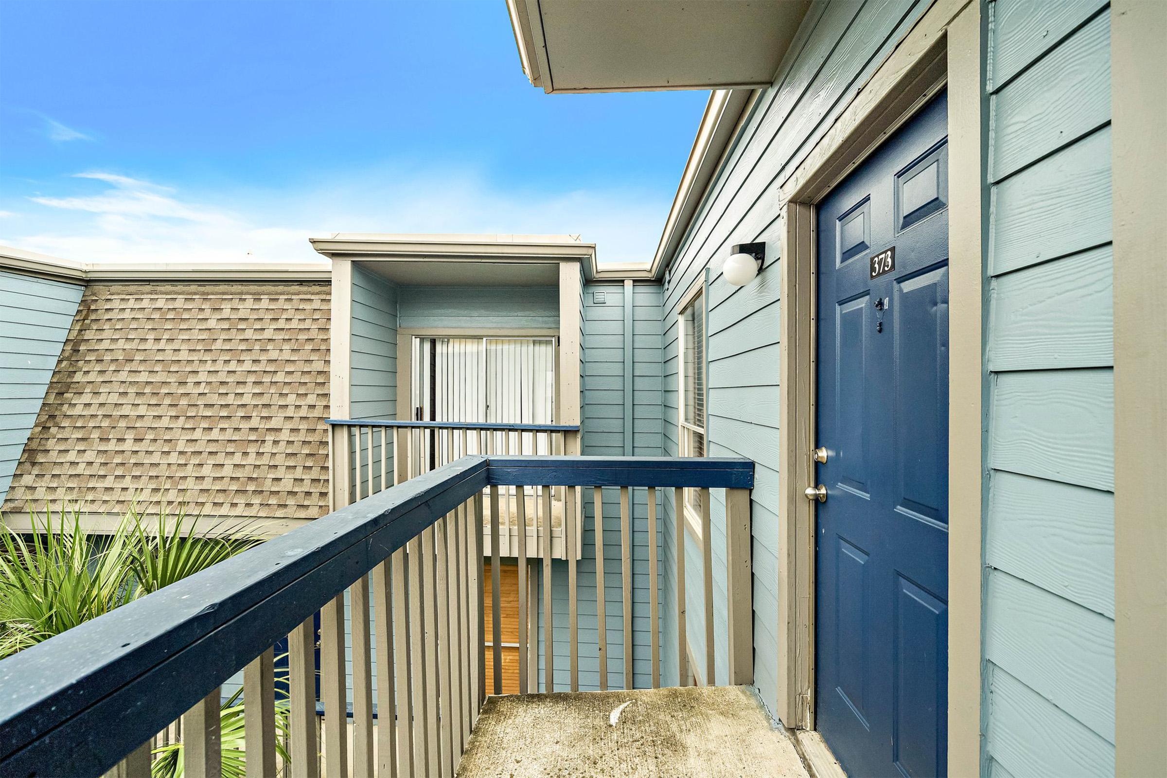 Exterior view of a multi-unit building showcasing a blue door with the number 873, surrounded by light blue siding and a balcony. A textured roof and palm leaves are visible in the foreground, with a clear blue sky in the background.