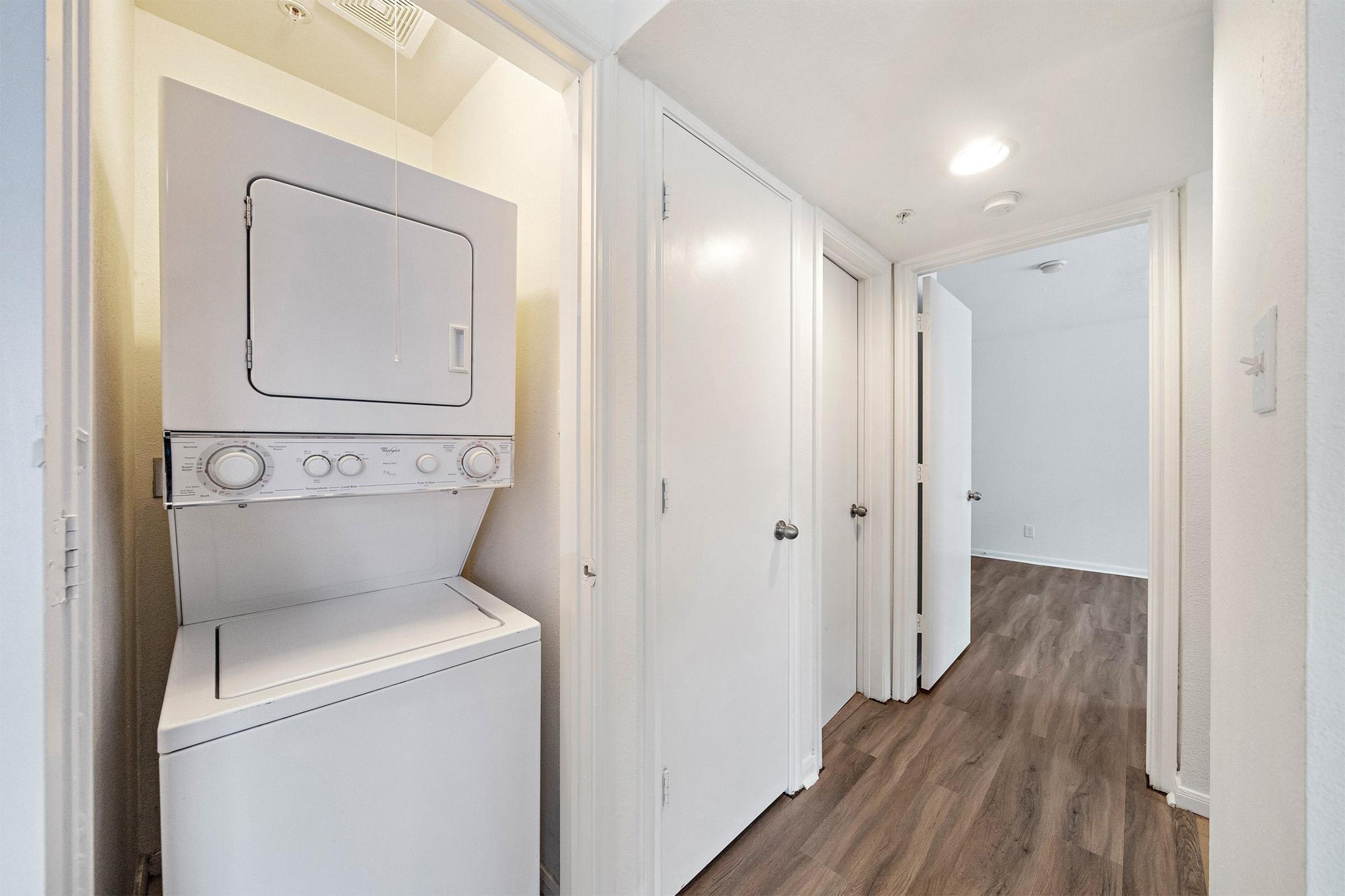A hallway with a stacked washing machine and dryer on the left. Light-colored walls and doors lead to other rooms. The floor is finished with a modern wood-like laminate. Natural light illuminates the space, giving it a bright and airy feel.