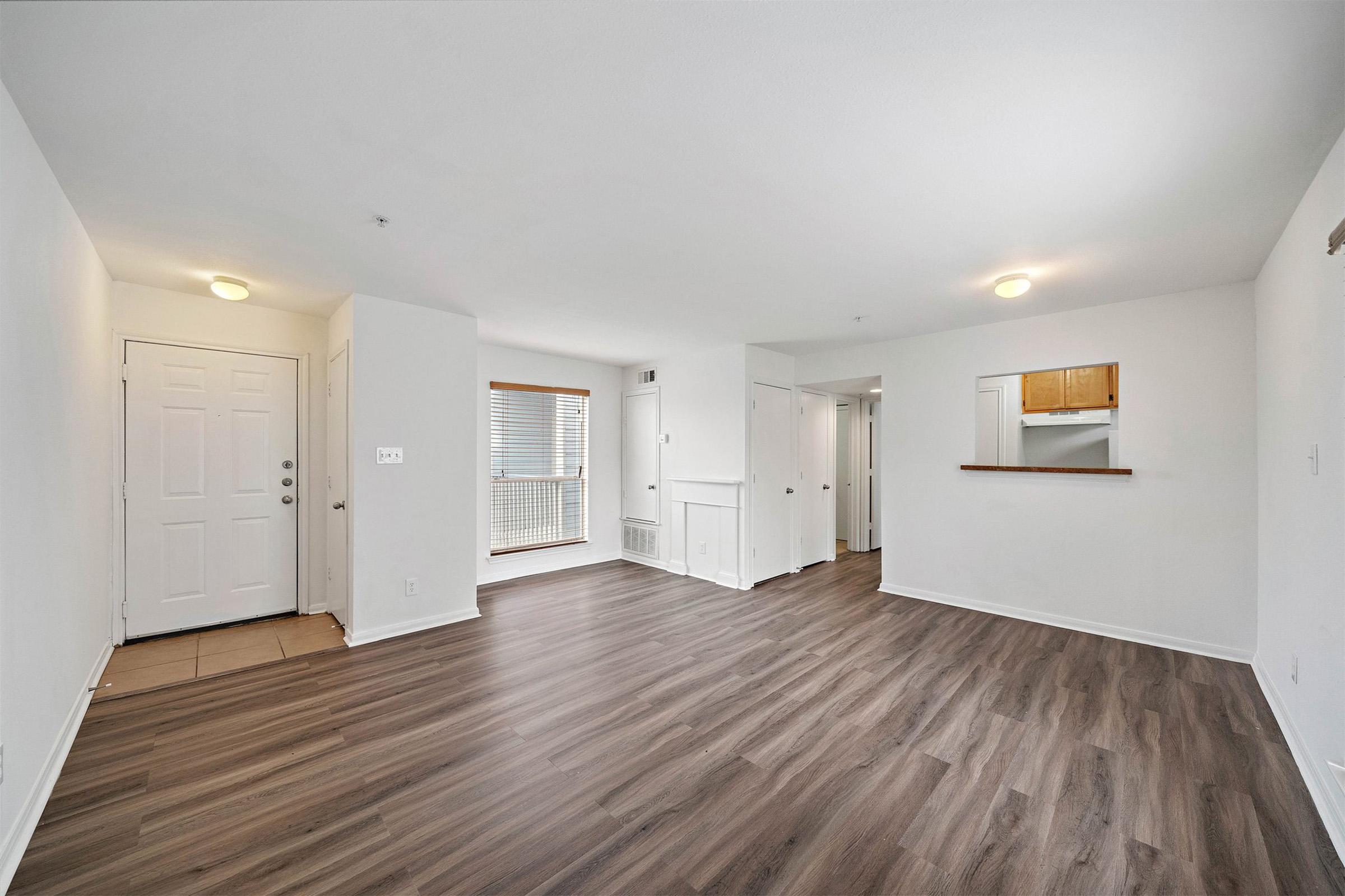 A spacious, well-lit living area with laminate flooring, featuring a front door, large windows with blinds, and an open layout that connects to a small kitchen area in the background. The walls are painted white, creating a clean and modern atmosphere.