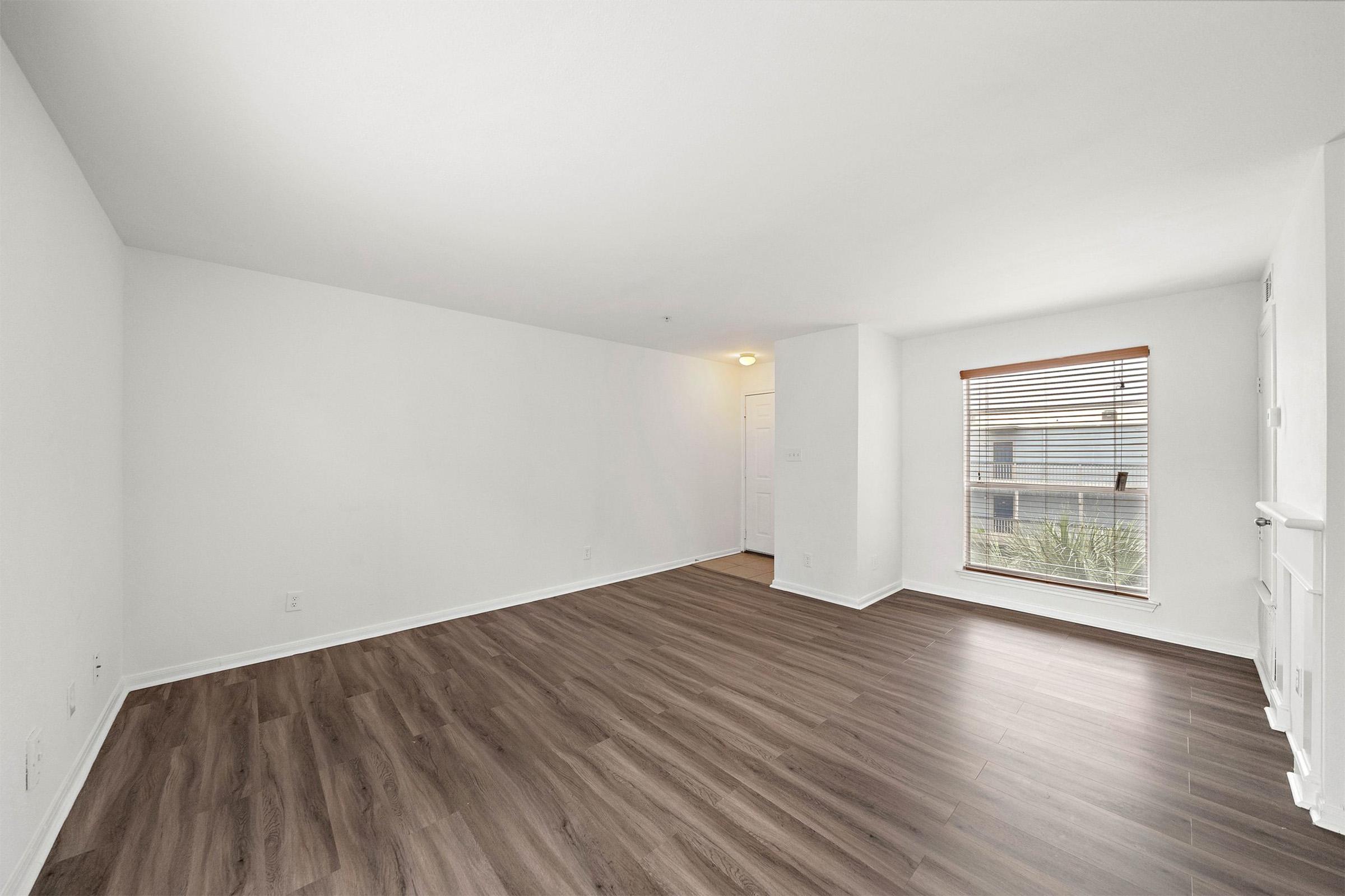 A spacious, empty room with light-colored walls and a brown laminate floor. A single window with wooden blinds allows natural light to enter. There is a door visible in the background leading to another space, and the overall atmosphere is clean and bright, suggesting potential for creative use.