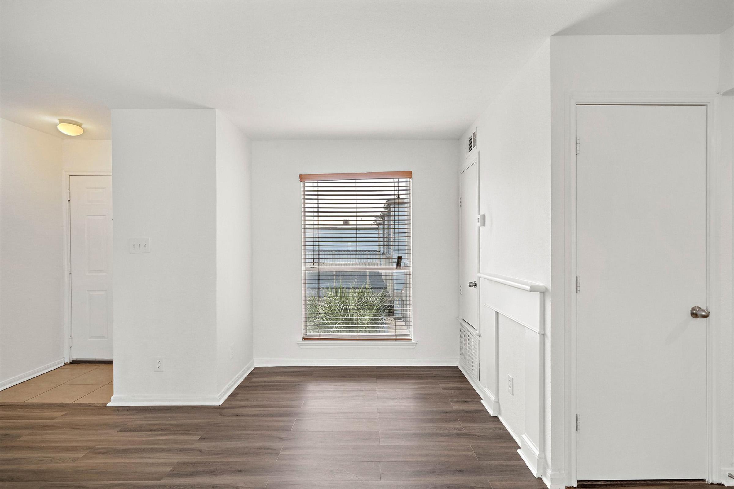 A bright, empty room with light-colored walls and a large window covered by closed blinds. The floor is a modern dark wood laminate. There is a door on the right and another visible door leading to a hallway on the left. The space appears clean and inviting, suitable for various uses.