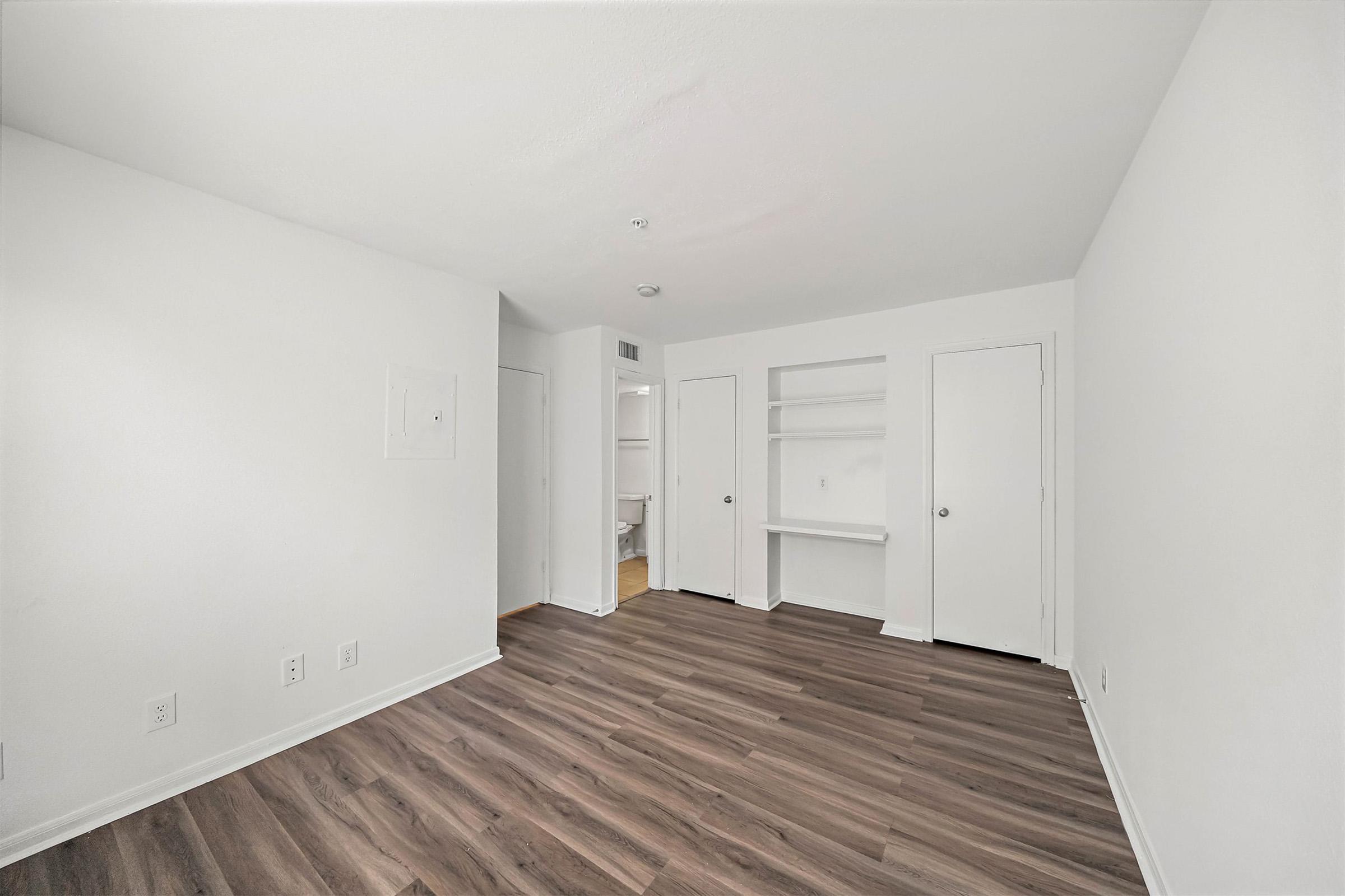 A vacant, modern room with white walls and a light brown wooden floor. It features a closet with sliding doors, a small built-in shelf, and two additional doors leading to different spaces. The room is well-lit and spacious, ready for furnishing.