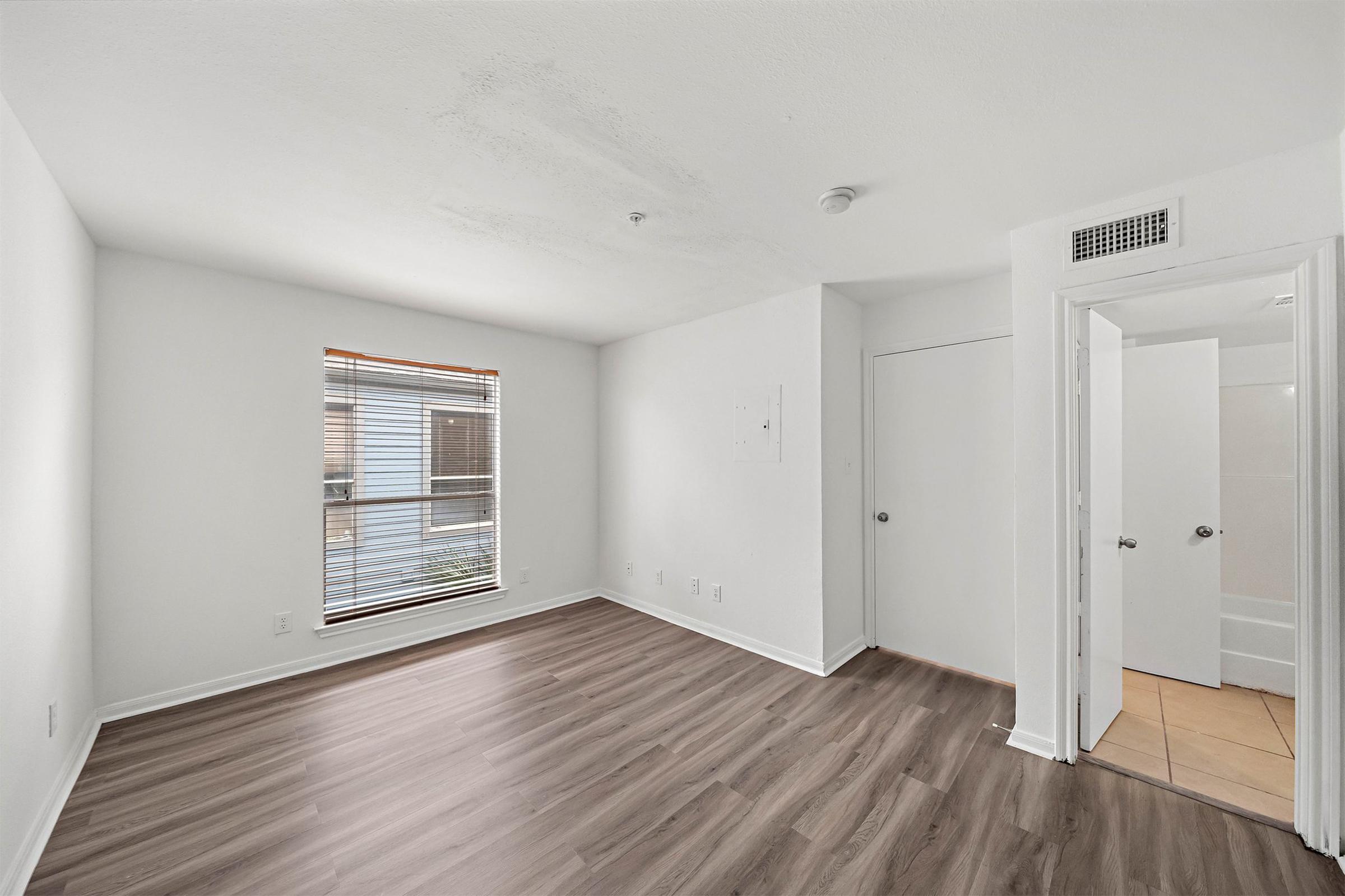 Empty room with light gray walls and a window featuring blinds. The floor is hardwood, and there is a door leading to a bathroom on the right side. The room has a clean, modern look with no furniture or decorations.