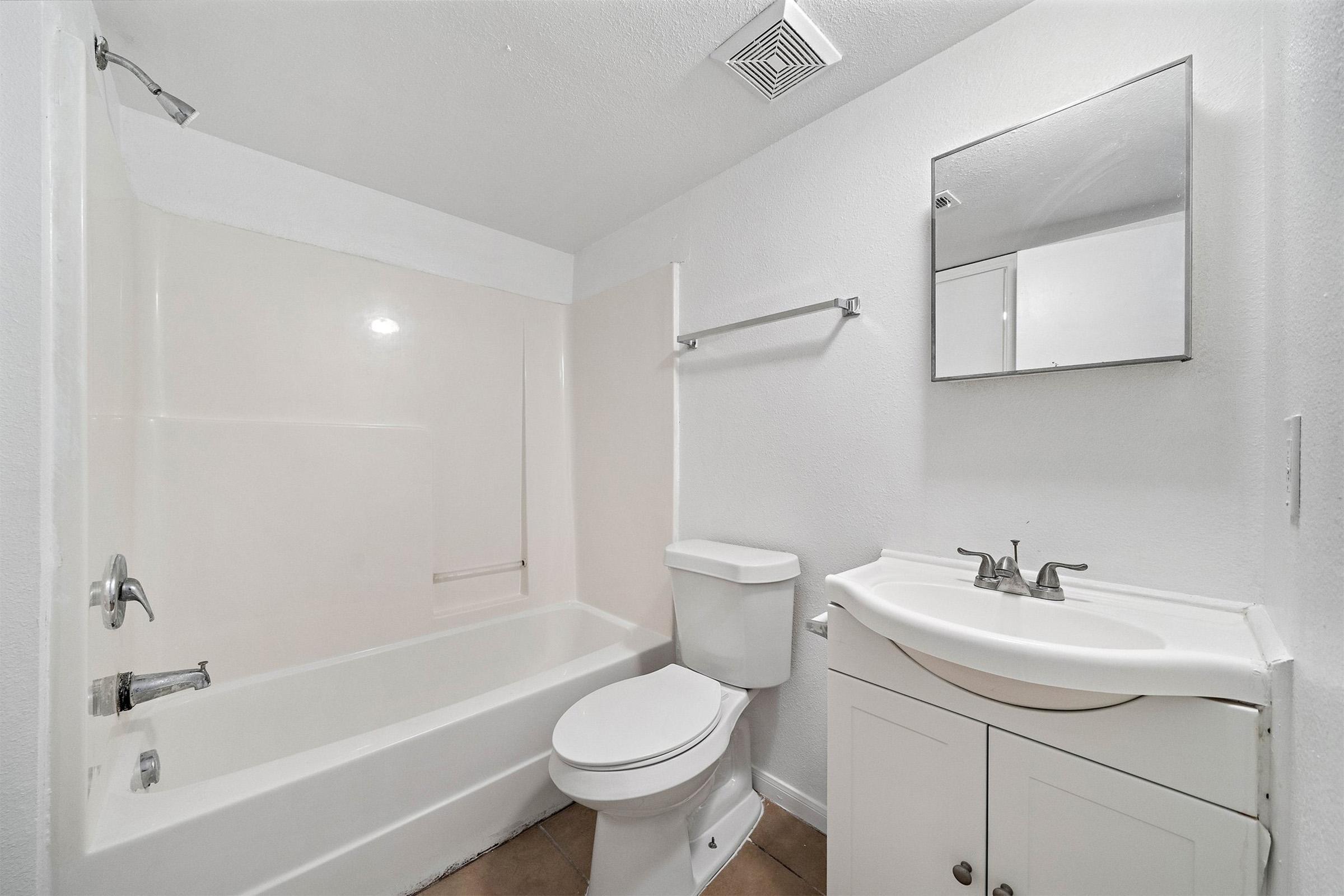 A clean and minimalistic bathroom featuring a white bathtub with a shower, a toilet, and a small sink with a mirror above it. The walls are painted white, and the floor is tiled in a light color, creating a bright and open space.