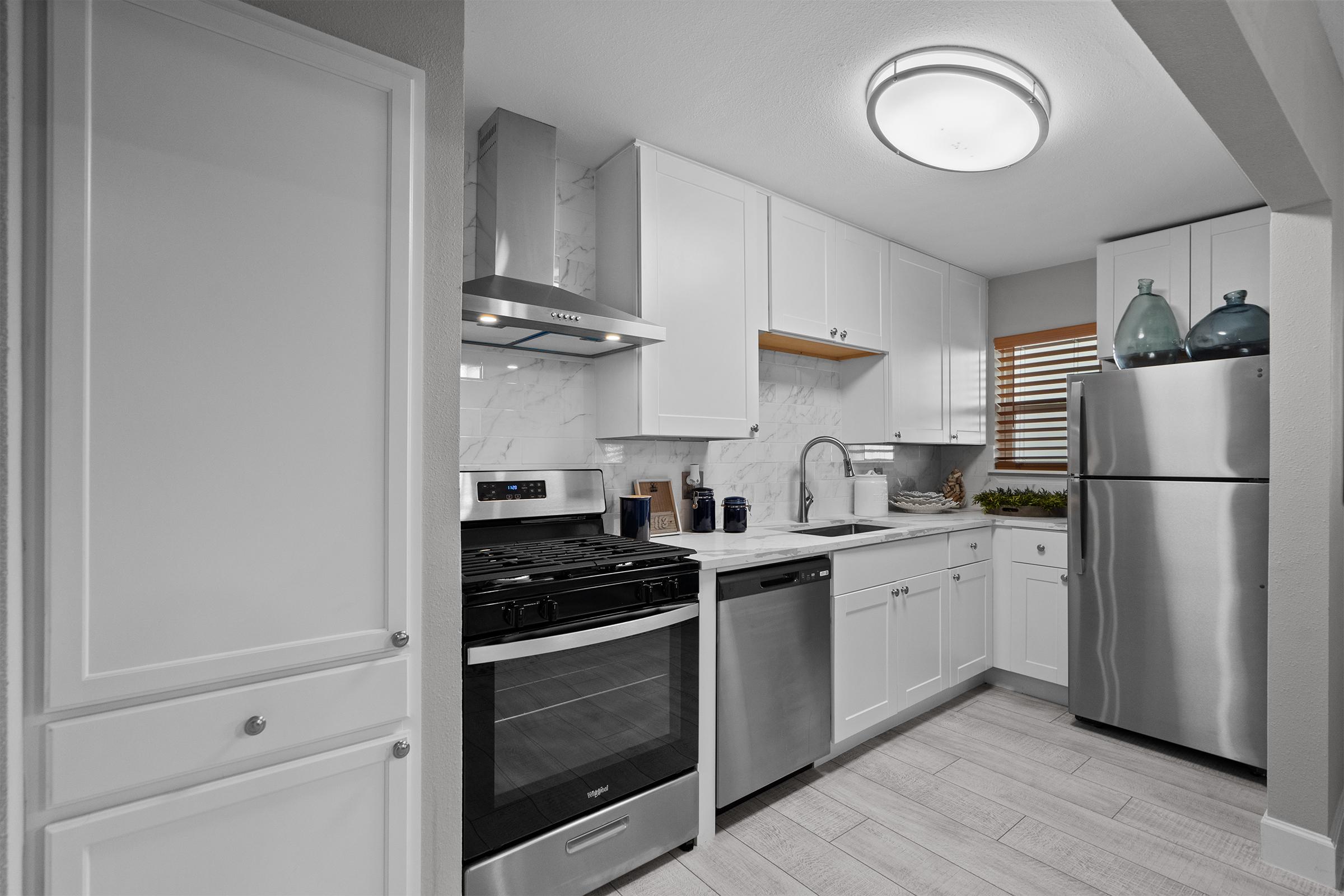 Modern kitchen featuring white cabinets, stainless steel appliances including a gas stove, refrigerator, and dishwasher. The countertop is light-colored with a sink, and there are decorative items on the shelves and window sill. A circular ceiling light illuminates the space.