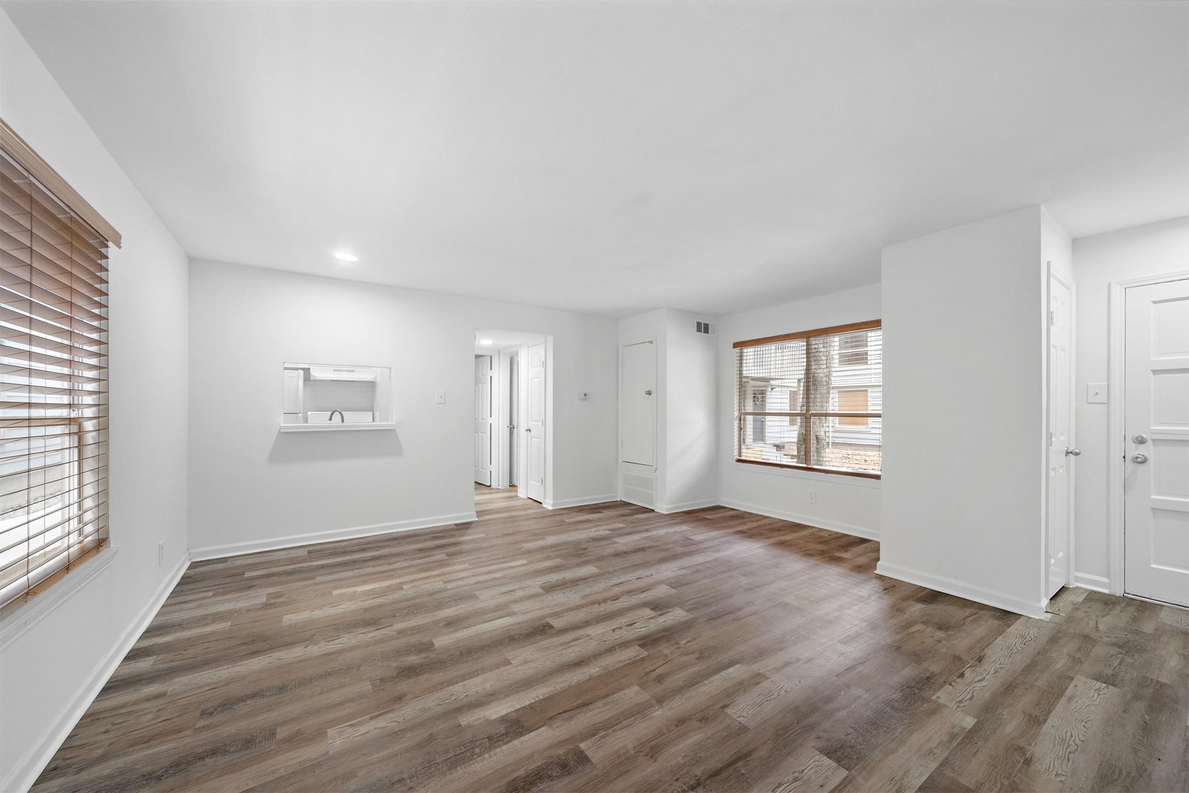 A spacious, well-lit room with light-colored walls, wooden floors, and large windows featuring blinds. There is a small mirror mounted on the wall and a doorway leading to another room. The overall aesthetic is modern and inviting, suitable for living or dining space.