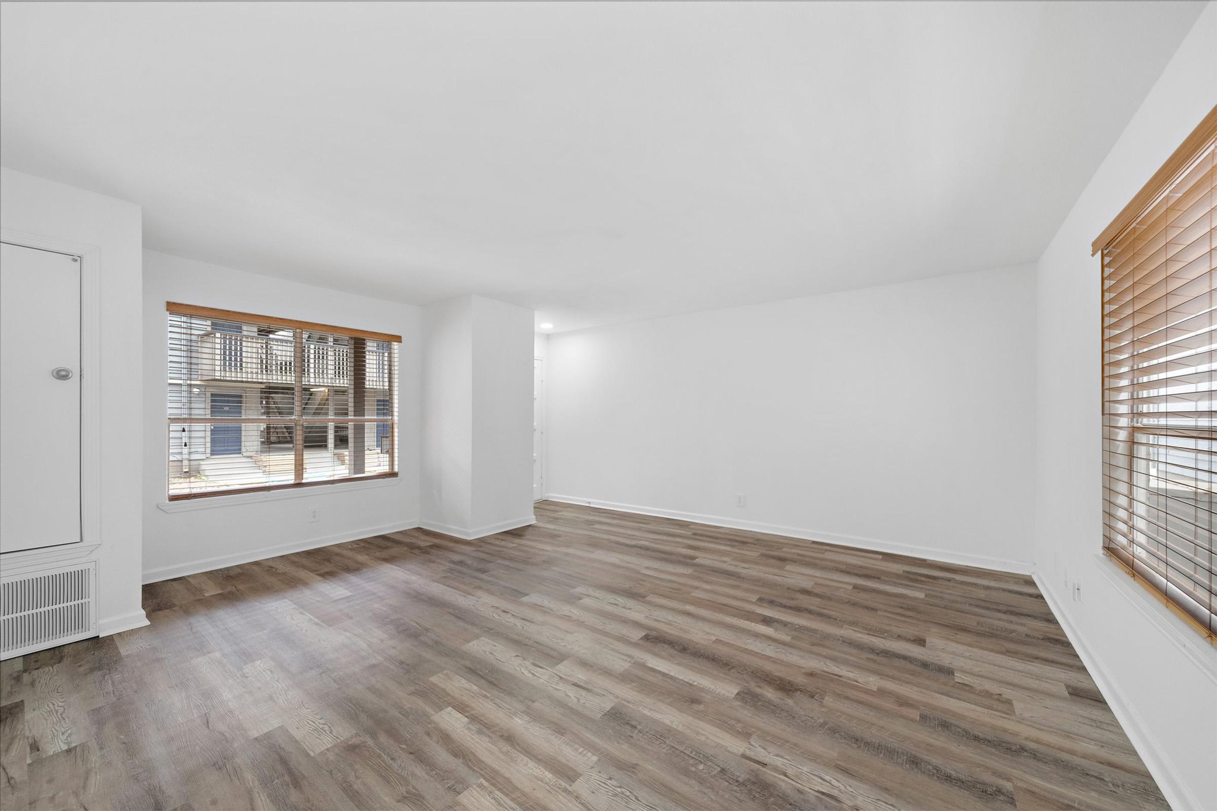 A spacious, empty room with light-colored walls and hardwood flooring. Large windows with wooden blinds allow natural light to fill the space. The room is brightly lit and appears clean and well-maintained, providing a blank canvas for potential furnishings and decoration.