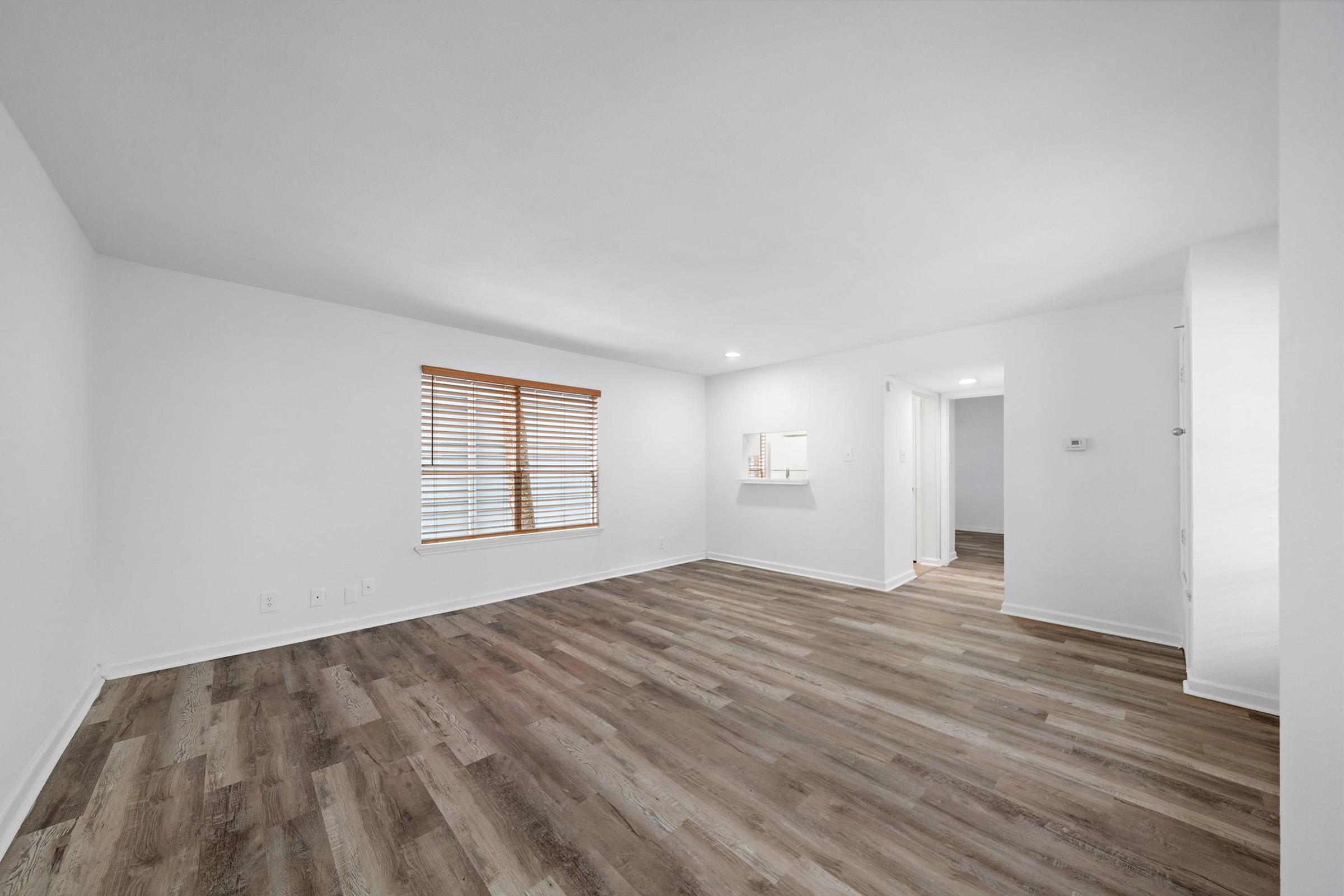 A vacant room with light-colored walls and wooden flooring. It features a large window with wooden blinds allowing natural light. A small opening leads to another area, possibly a kitchen, and there are doors to adjacent spaces, contributing to an open, airy layout.
