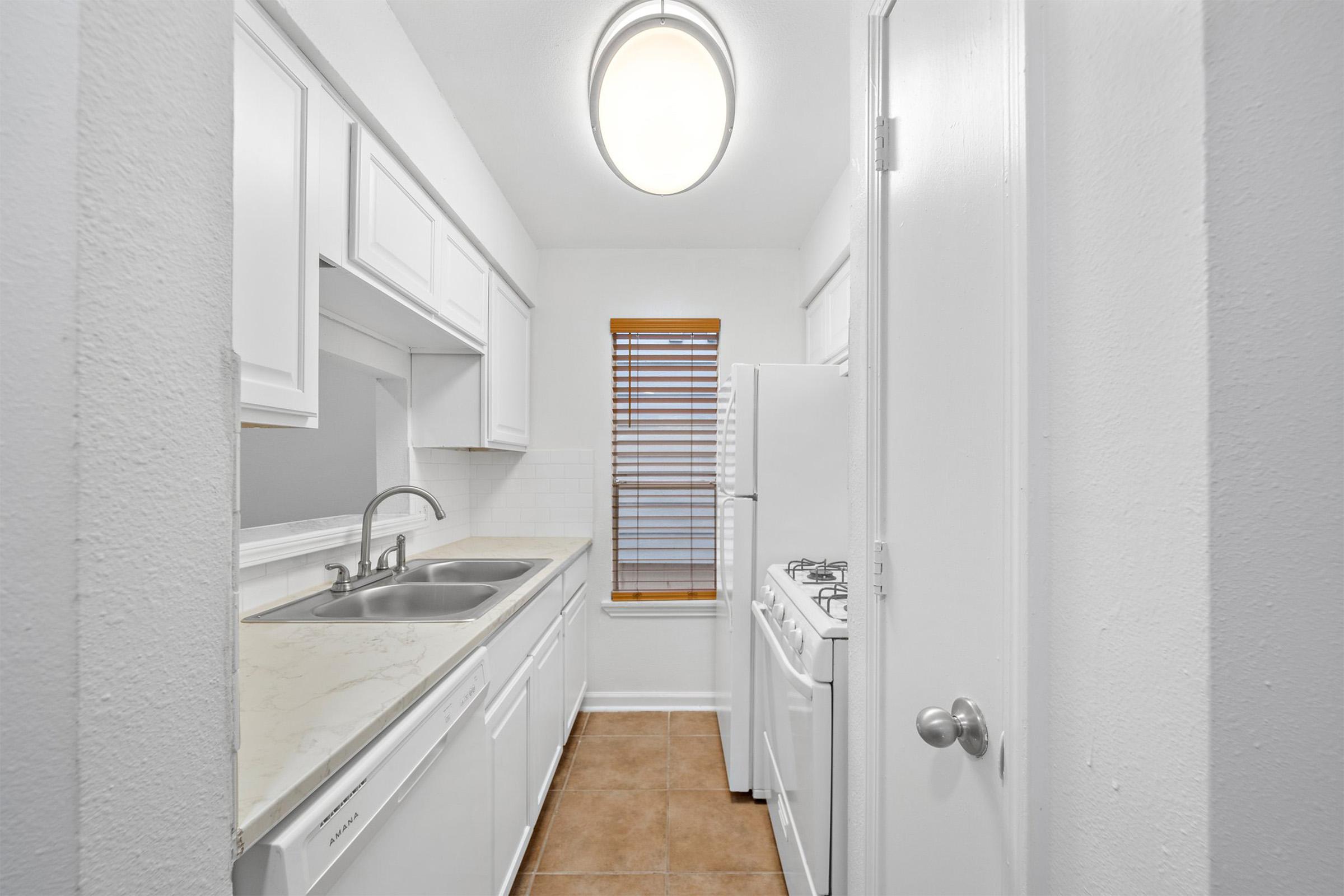 A narrow kitchen featuring white cabinets, a double sink, a gas stove, and a refrigerator. The room is brightly lit by a ceiling light and a window with blinds, allowing natural light to enter. The walls are painted white, and the floor is tiled with beige tiles.