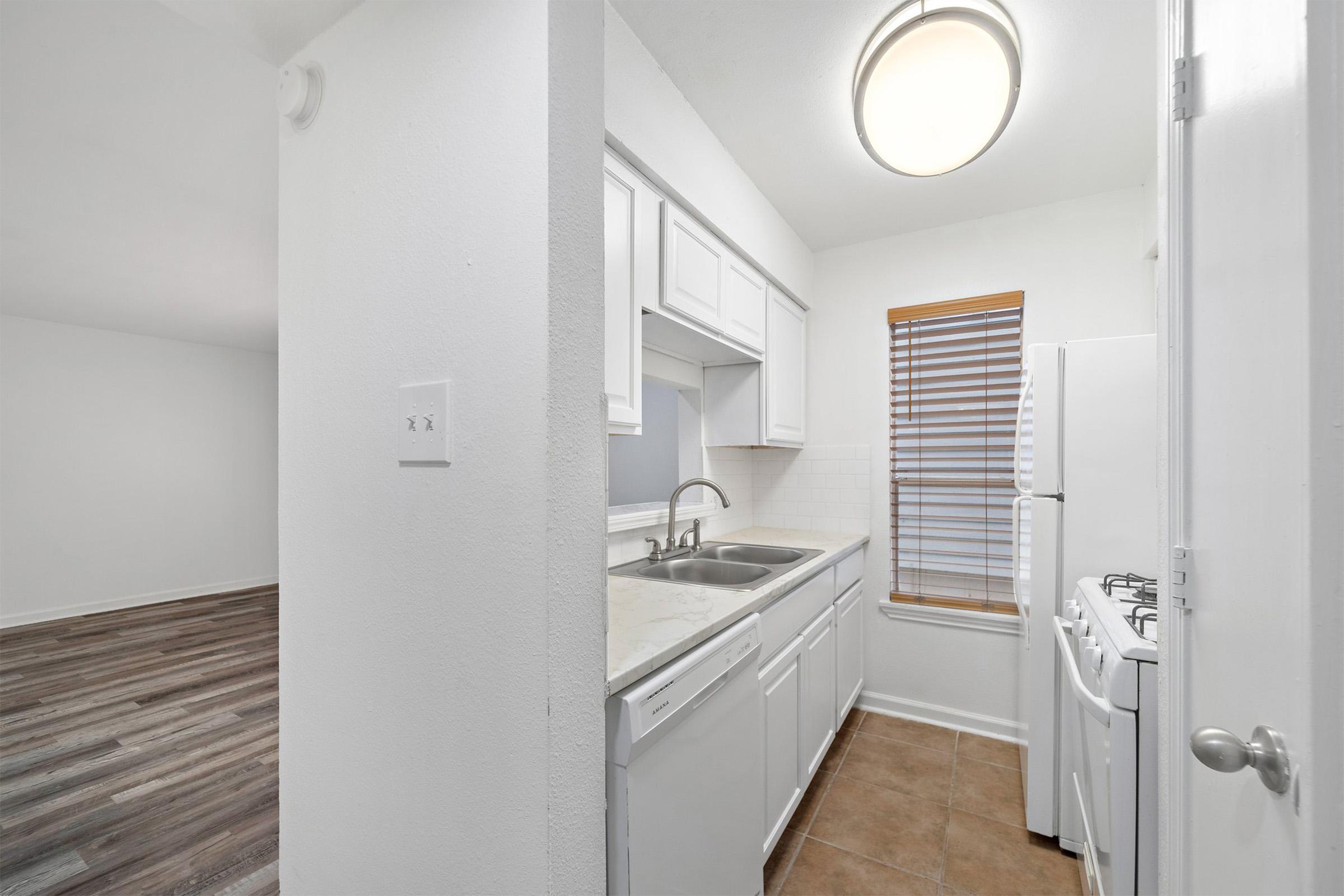 A clean, modern kitchen featuring white cabinetry, a double sink, and white appliances including a refrigerator and stove. The area has a window with wooden blinds, allowing natural light to enter. The flooring transitions to a wooden style in the adjacent room, which is not visible in this section.