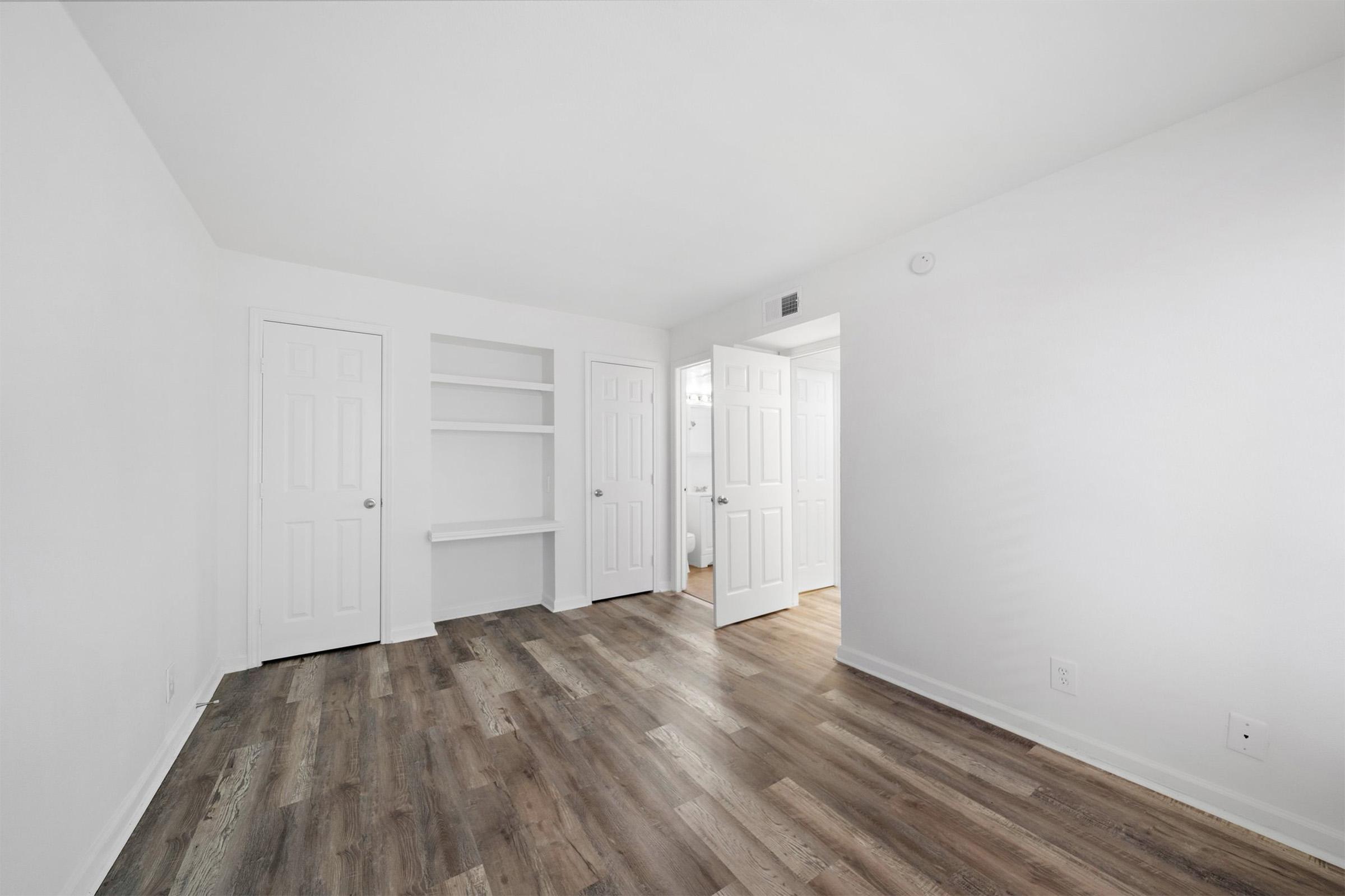 A bright, empty room with white walls and a light wood floor. Two closed doors are on the left, leading to closets or storage, while a third door on the right leads to another room. There is a small shelf built into the wall. The space is well-lit, creating a clean and open atmosphere.