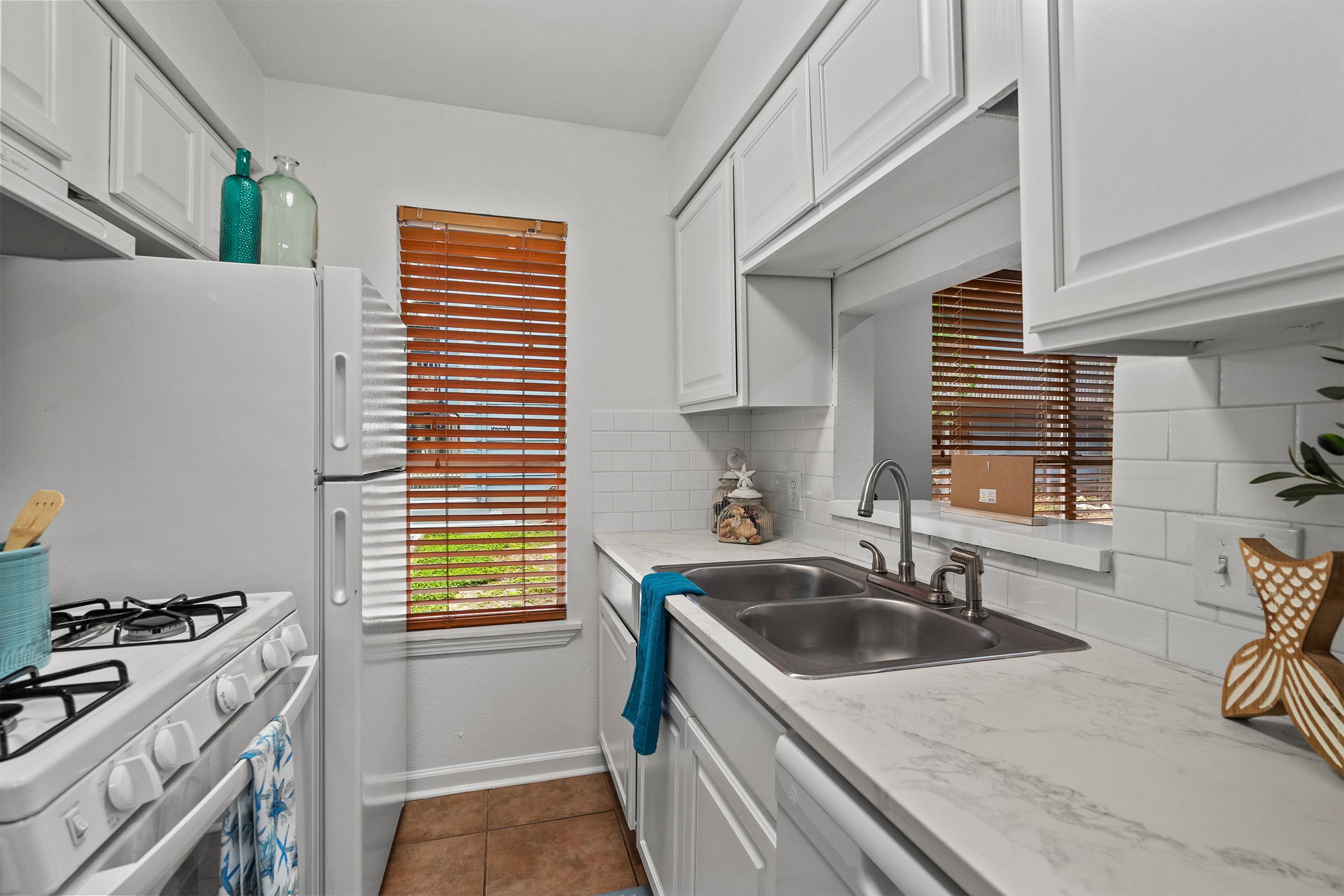 A clean, modern kitchen featuring white cabinetry, a gas stove, and a double sink with a marble countertop. Sunlight streams in through two windows with wooden blinds, illuminating decorative elements like a turquoise dish and jars. A towel hangs neatly from the stove, adding a touch of color.