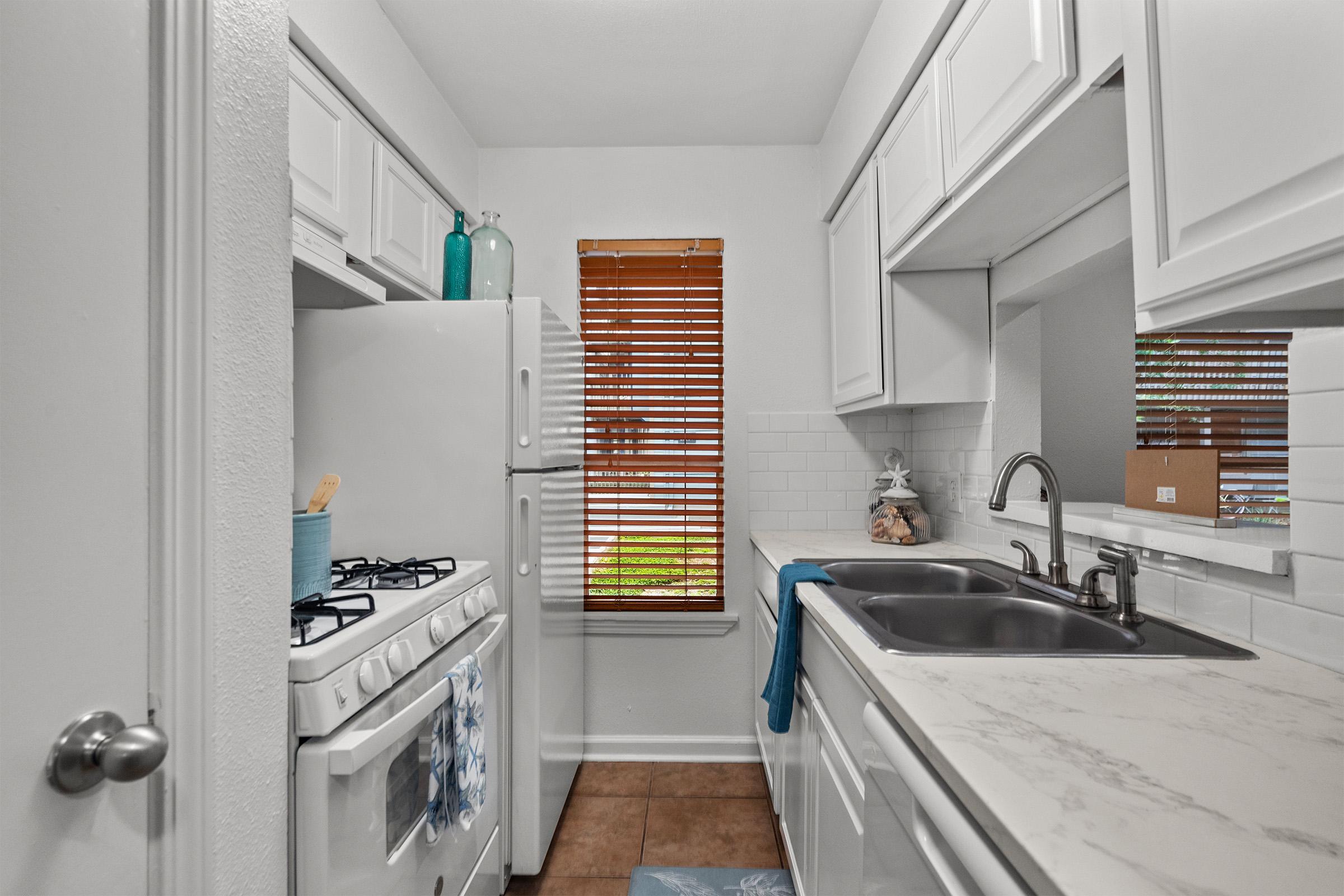 A small modern kitchen featuring white cabinets and a marble countertop. There's a gas stove on the left, a double sink with a blue towel, and a large window with wooden blinds allowing natural light to enter. A few decorative items are placed on the countertop and window sill.