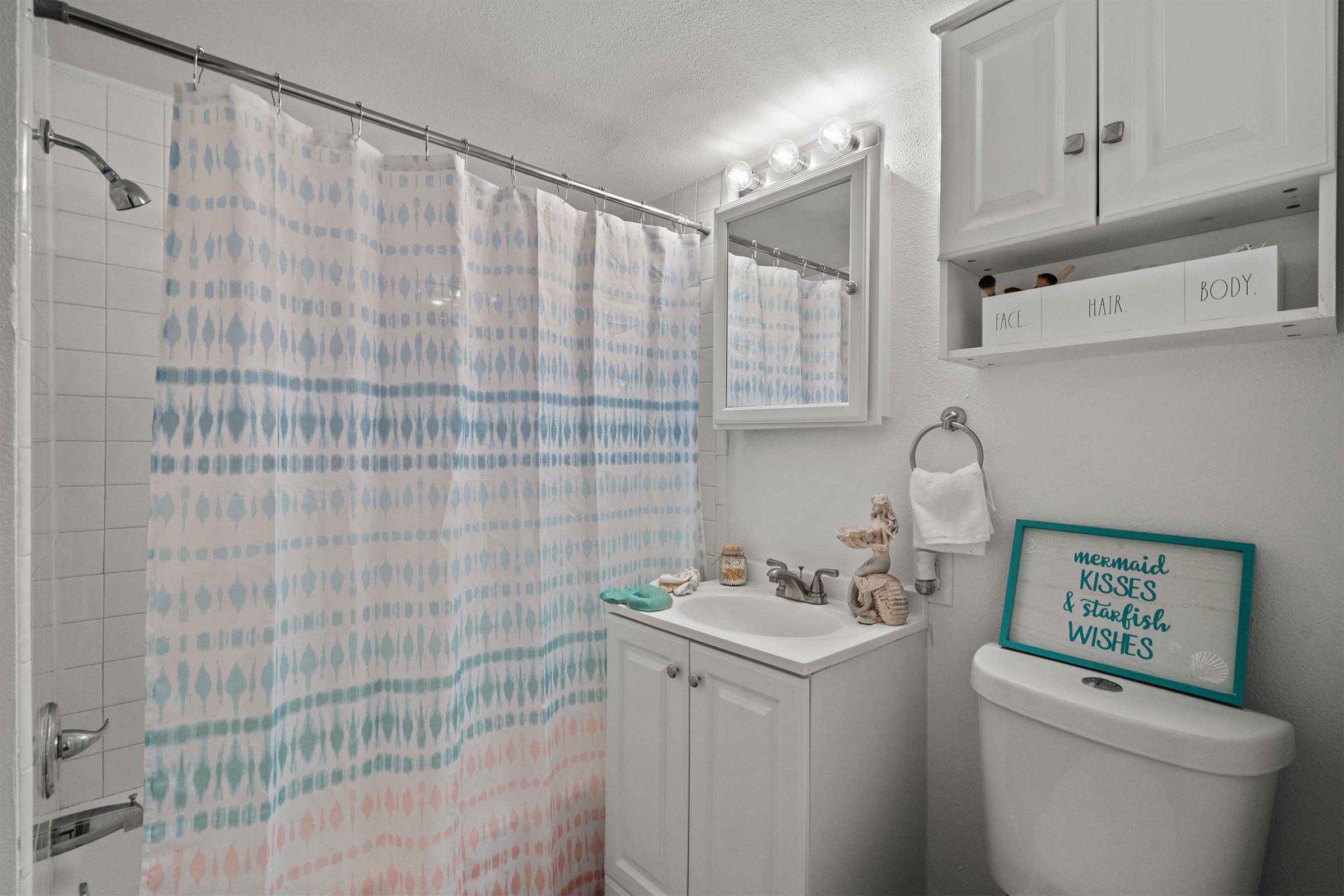 A bright bathroom featuring a pastel-colored shower curtain with a tie-dye pattern. It includes a small white cabinet above the toilet, a mirror with lights, and decorative items like a sign that reads "mermaid KISSES & starfish WISHES." The space is clean and thoughtfully arranged, creating a welcoming atmosphere.