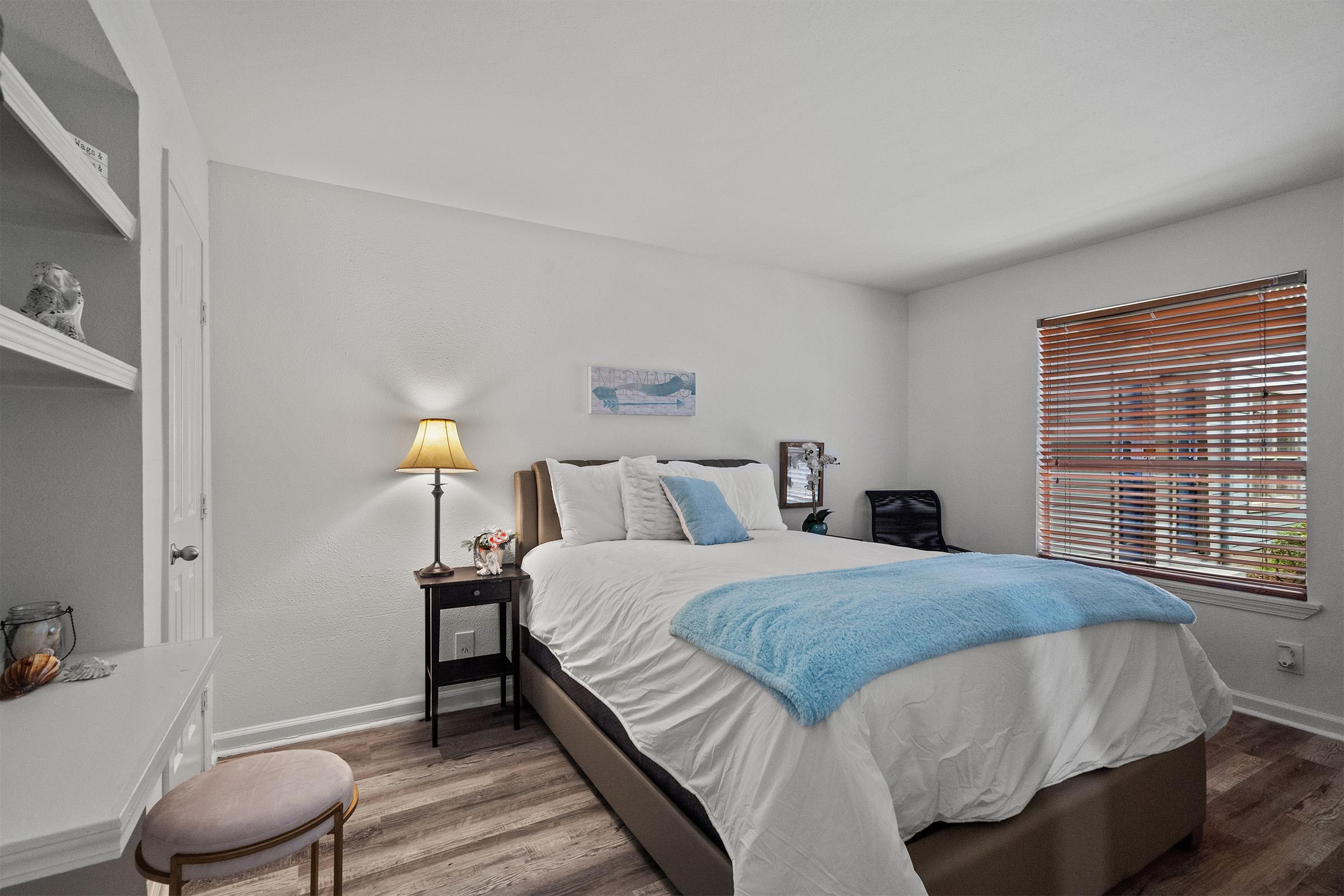 Cozy bedroom featuring a large bed with a blue throw blanket, a nightstand with a lamp, and a floral arrangement. A window with wooden blinds allows natural light to enter, illuminating the light-colored walls and wooden flooring. A small chair and a framed picture are also visible in the room.