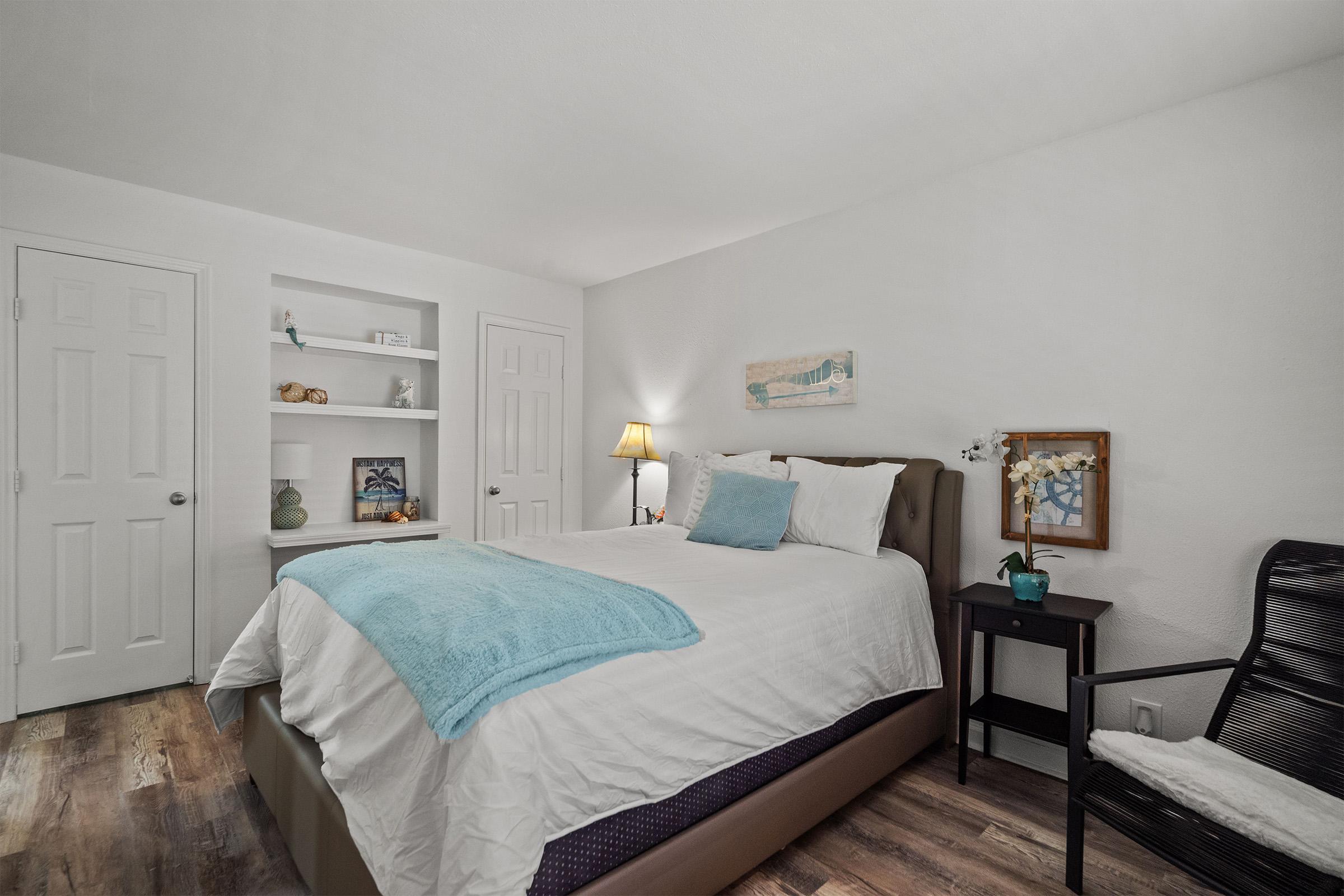 Cozy bedroom featuring a queen-sized bed with a light blue blanket, a small nightstand with a lamp, and a comfortable chair. The room is decorated with a piece of artwork and has a shelf displaying decorative items. White walls and wood-like flooring create a warm and inviting atmosphere.