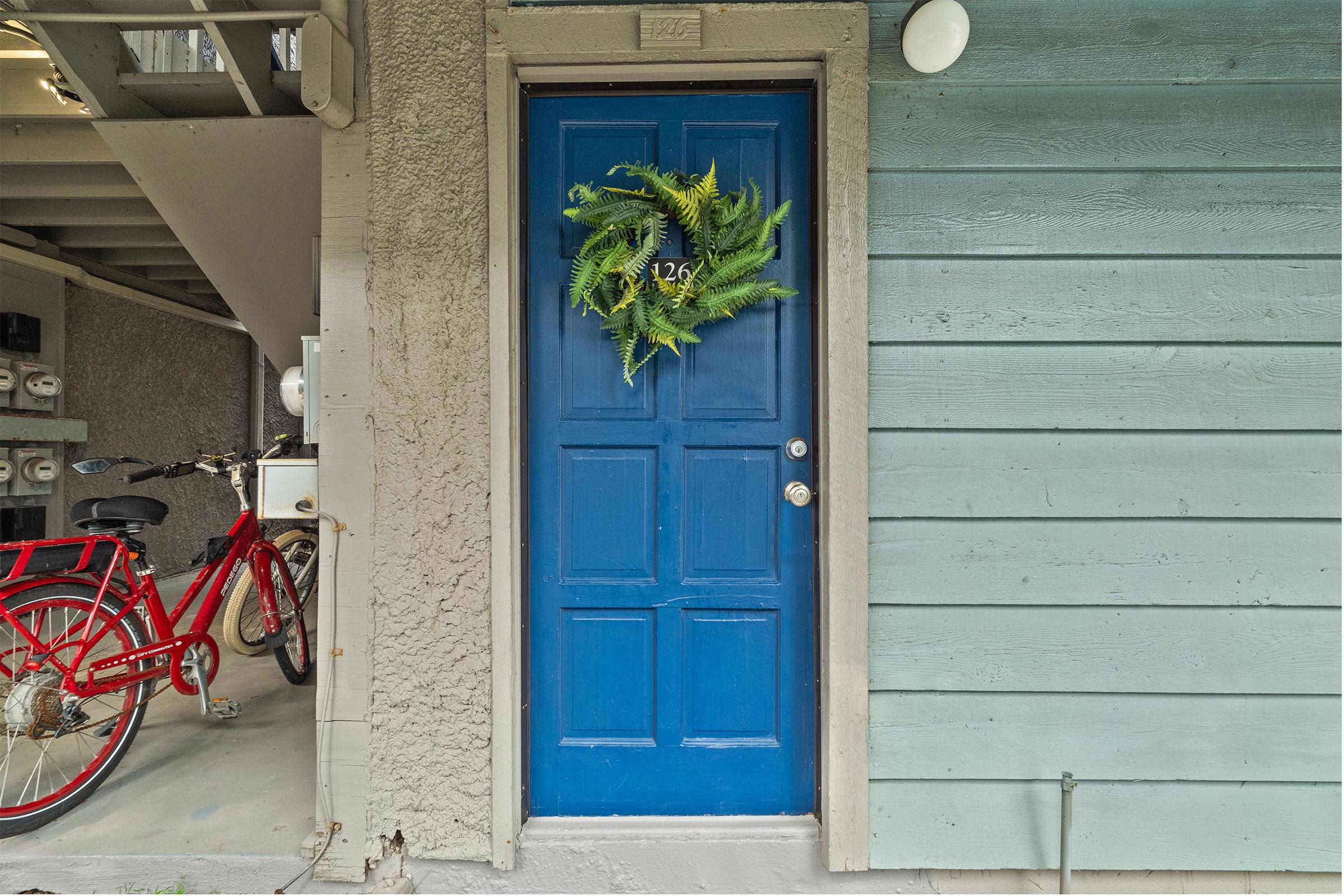 A blue front door with a green wreath hangs on it, located on a house with light blue siding. To the left, a red bicycle leans against the wall, and there is visible parking and utility area behind the door. The entrance has a simple and welcoming appearance.