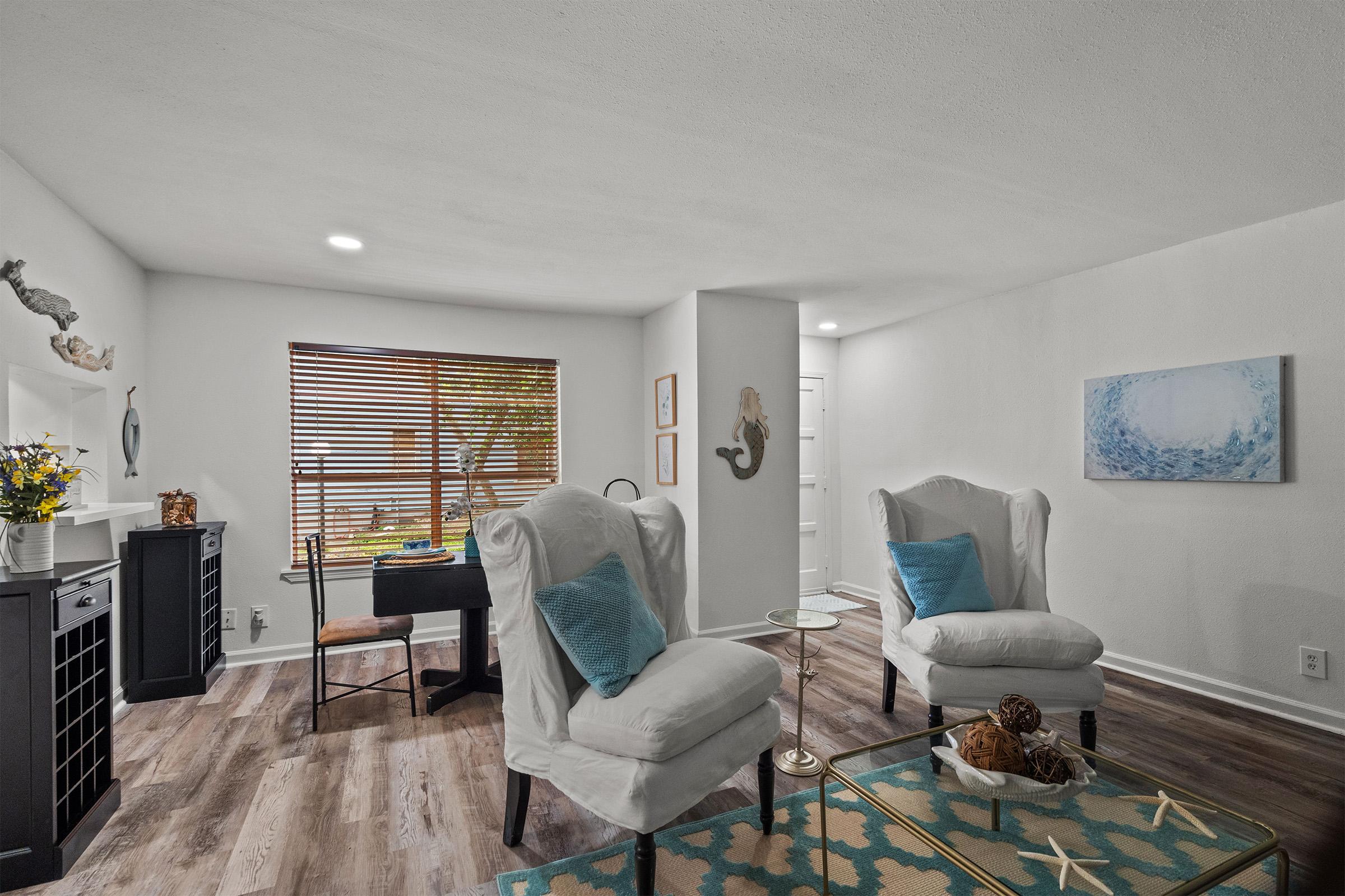 A bright, modern living room featuring two white upholstered chairs with blue pillows, a glass side table, and a decorative rug. Natural light filters through wooden blinds, illuminating a small desk and artwork on the walls, creating a cozy and inviting atmosphere.