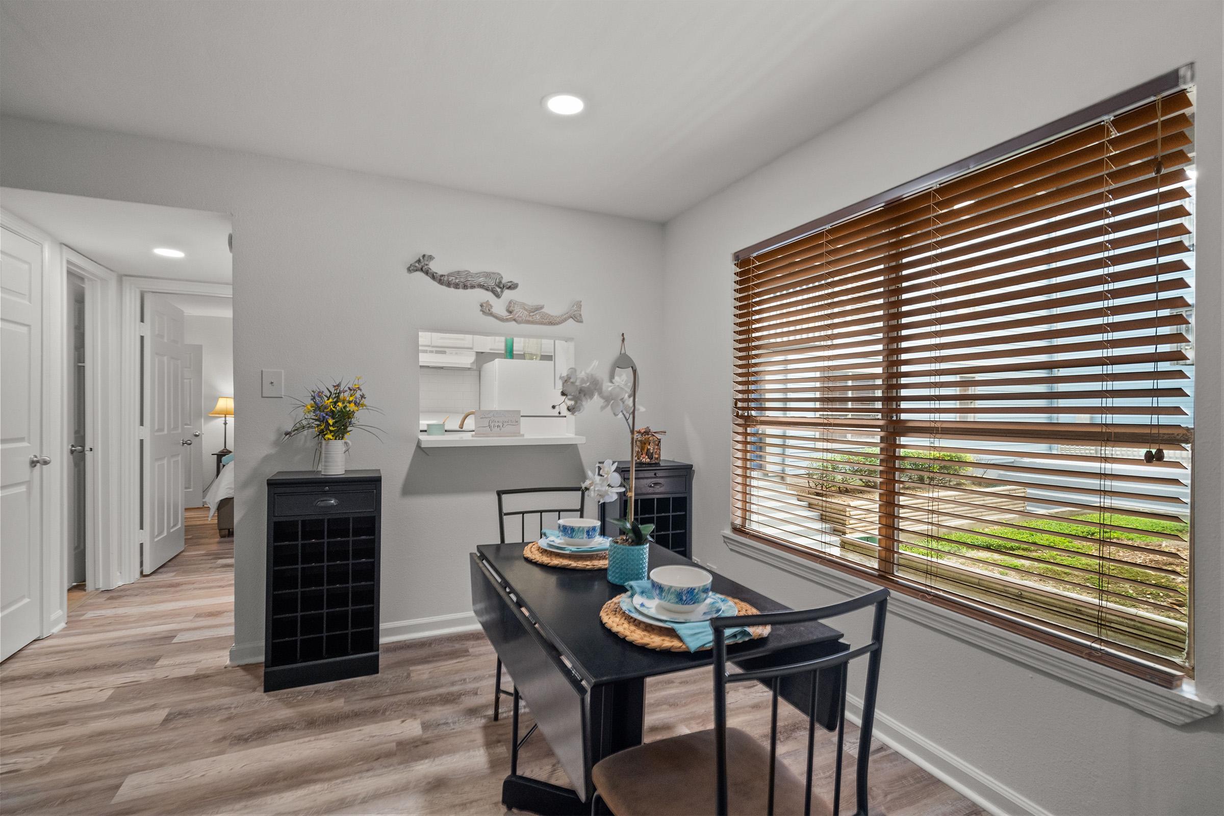 A cozy dining area featuring a black table set for two, with blue dishes and placemats. A window with wooden blinds allows natural light, highlighting the room's neutral decor. A small decorative shelf with a figurine and flowers adds charm, while a modern kitchen area is visible in the background.
