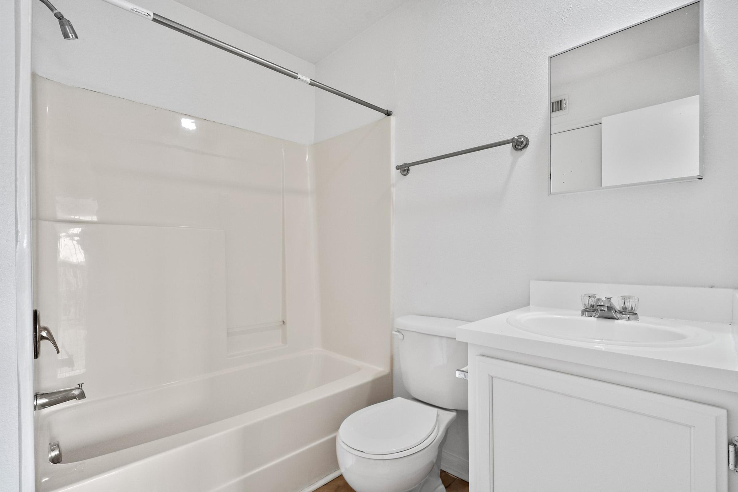 A clean and modern bathroom featuring a bathtub with a shower, a white toilet, and a simple vanity with a sink. A mirror is mounted above the sink, and the walls are painted white. The flooring is light-colored, creating a bright and spacious appearance.