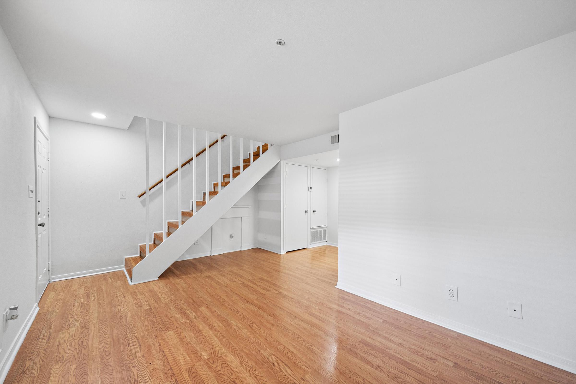 Empty interior of a room featuring a wooden staircase, light-colored walls, and hardwood flooring. A door is visible on the left, and there are white doors leading to other areas in the background. The space is well-lit and uncluttered, giving a sense of openness.