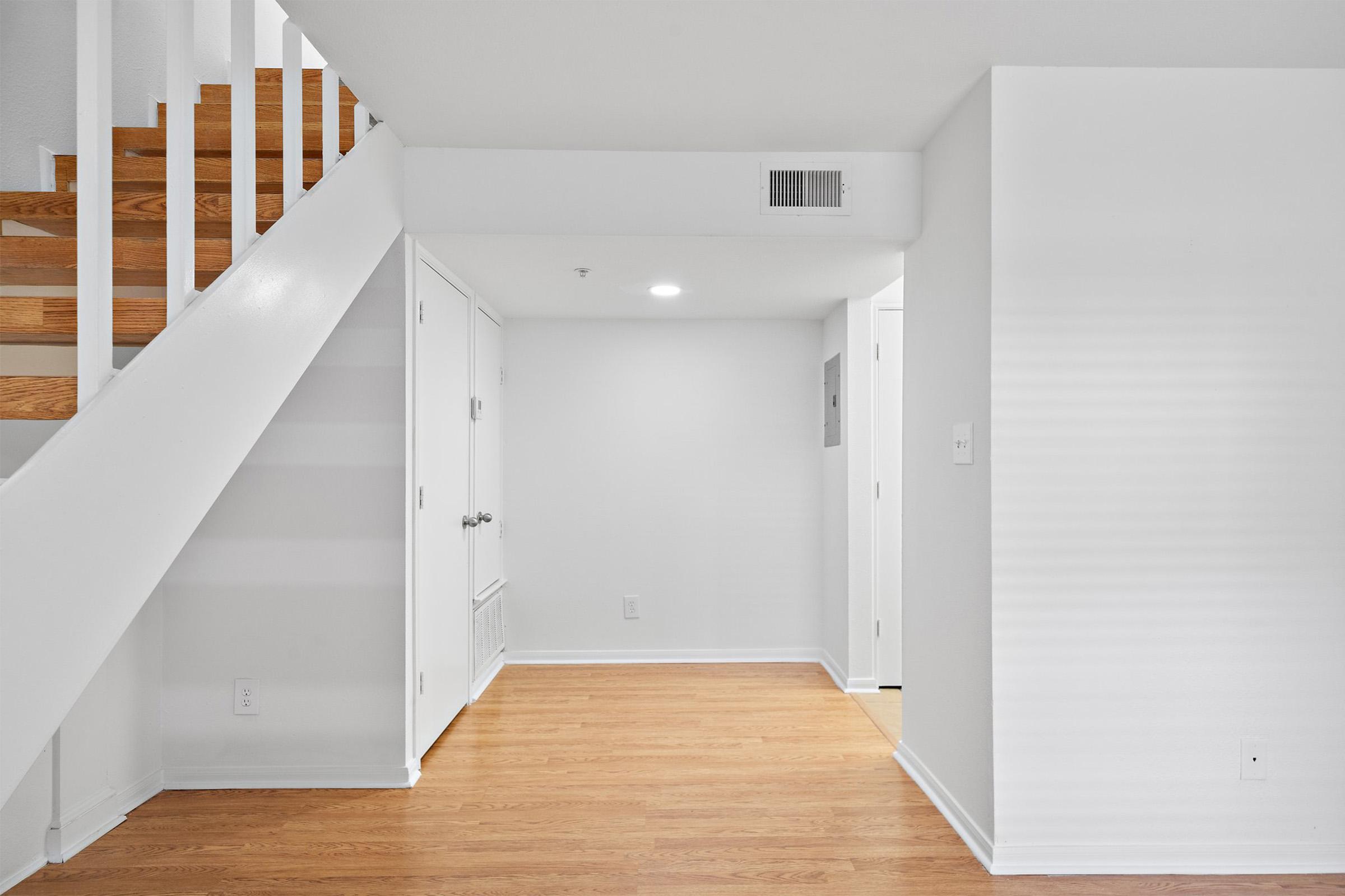 Interior view of a hallway with wooden stairs leading to the upper floor. Light-colored walls and smooth wooden flooring create a bright and airy atmosphere. A closed door and a small closet are visible, with a ceiling light illuminating the space. The design is modern and minimalistic.