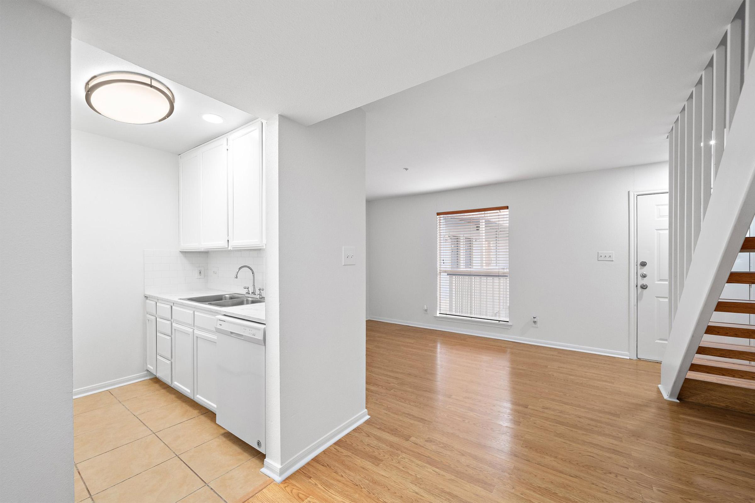 A modern interior space featuring a kitchen with white cabinets and a dishwasher, adjacent to a living area with hardwood floors and a large window. Stairs lead to the upper level, and the room is illuminated by a ceiling light. The walls and trim are painted in neutral tones, creating a bright and airy atmosphere.