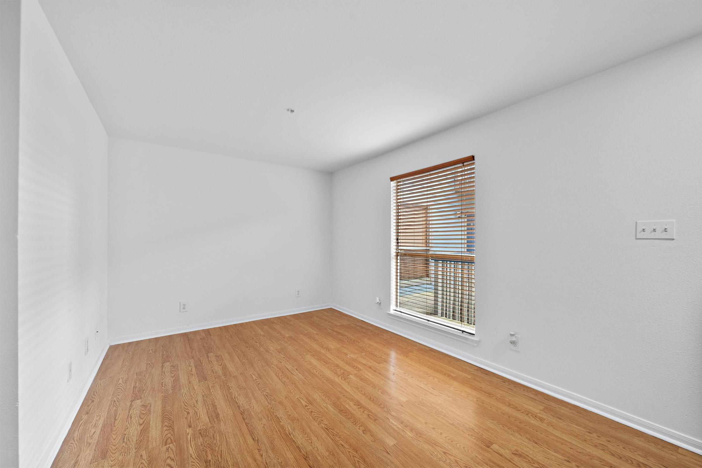 A well-lit, empty room with light hardwood flooring and white walls. A window with wooden blinds allows natural light to enter, casting soft shadows. The space appears clean and minimalistic, ideal for personal decoration or furnishing.