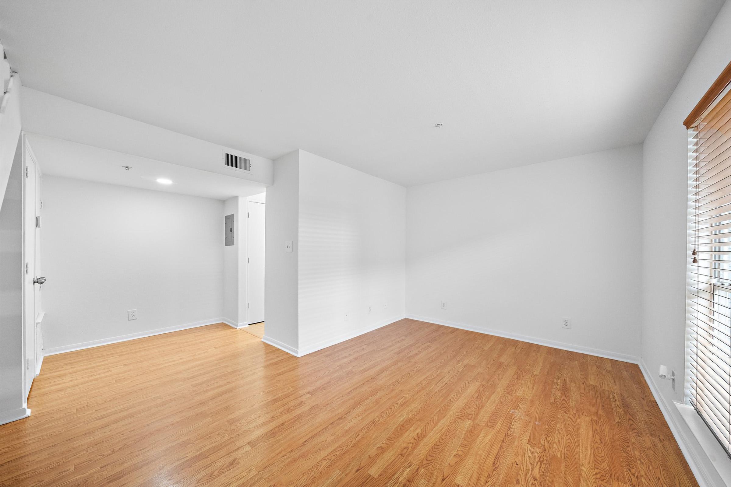 A spacious and bright room with light wooden flooring, white walls, and a large window with blinds. The room features an open layout with a doorway leading to another area. Natural light fills the space, creating a warm and inviting atmosphere.