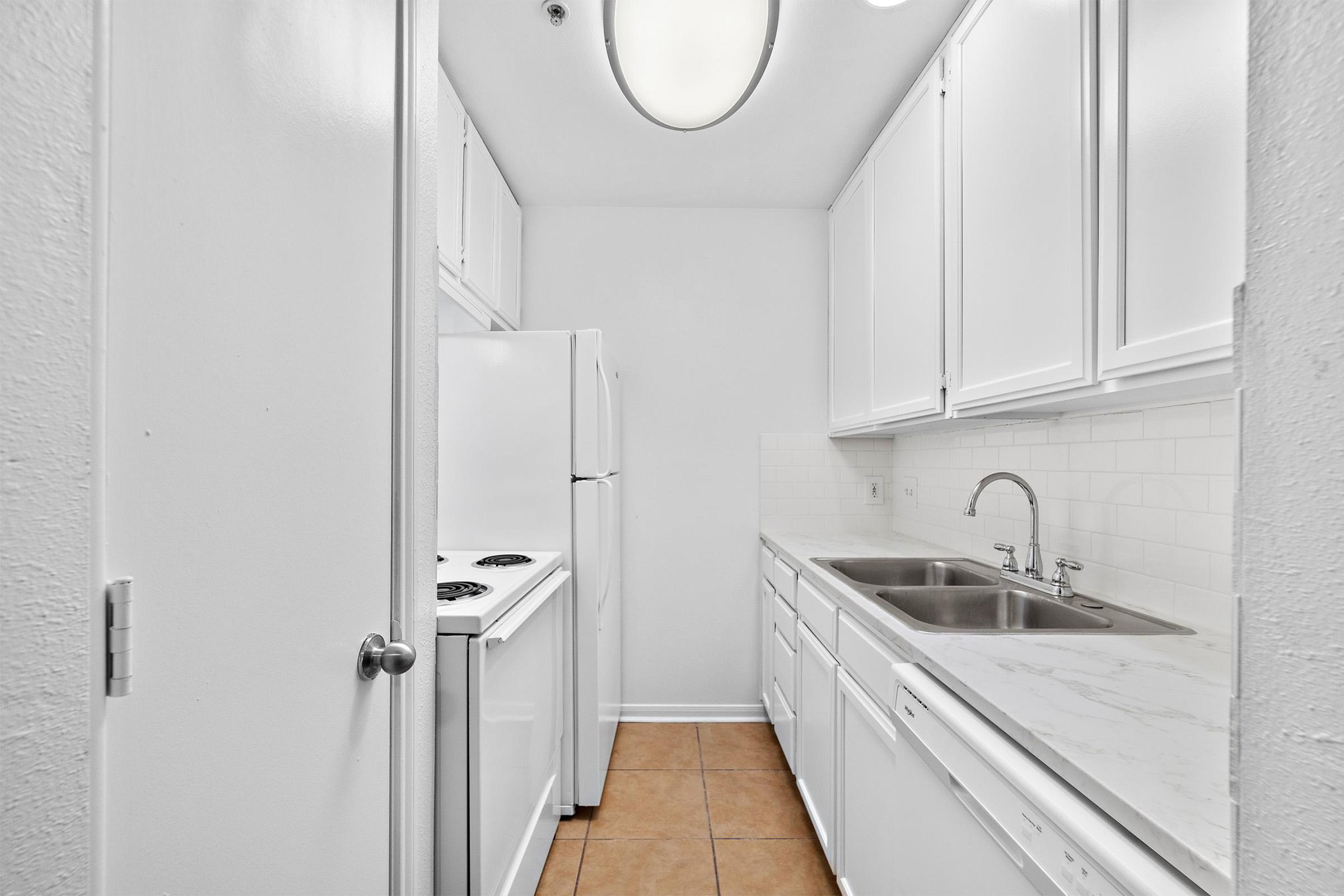 Narrow kitchen featuring white cabinetry, a double sink, and a two-burner stove. The space includes a white refrigerator and light-colored tile flooring, with soft overhead lighting illuminating the area. The walls are painted white, adding to the bright and clean appearance.