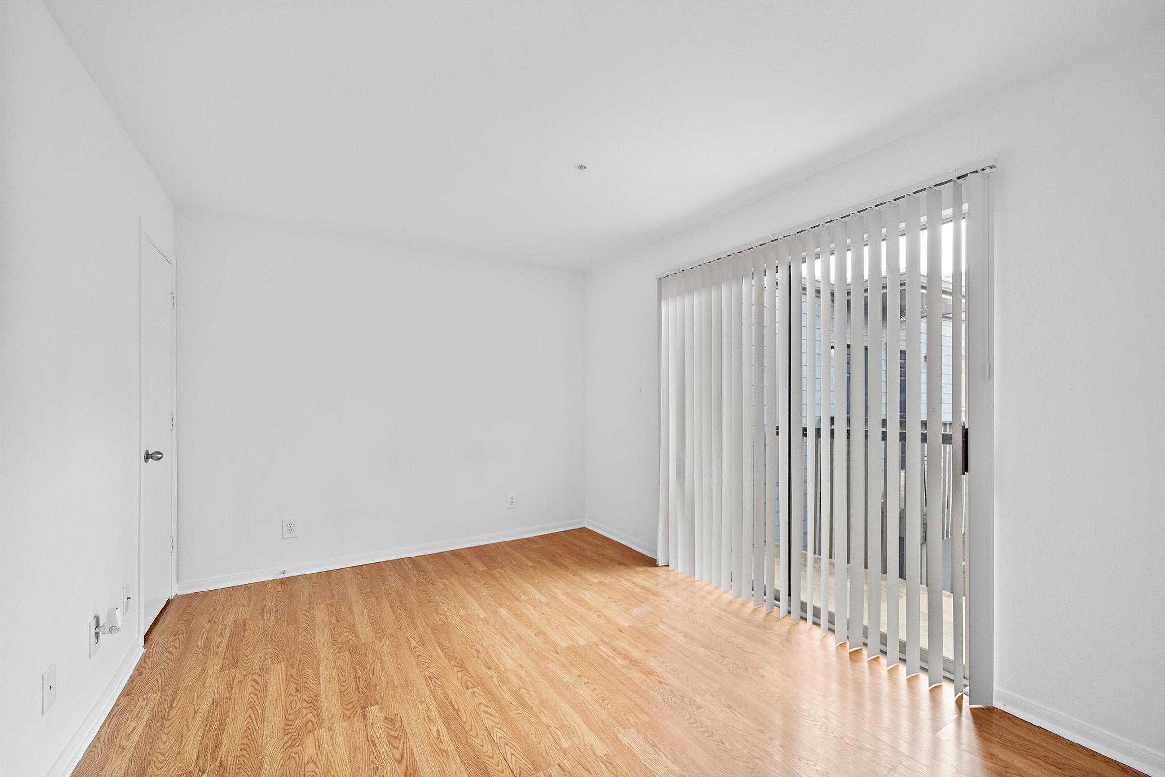 Empty room with light-colored walls and hardwood flooring. A large window with vertical blinds allows natural light to enter, and there is a door on the left side, leading to another area. The space appears clean and ready for decoration or furniture placement.