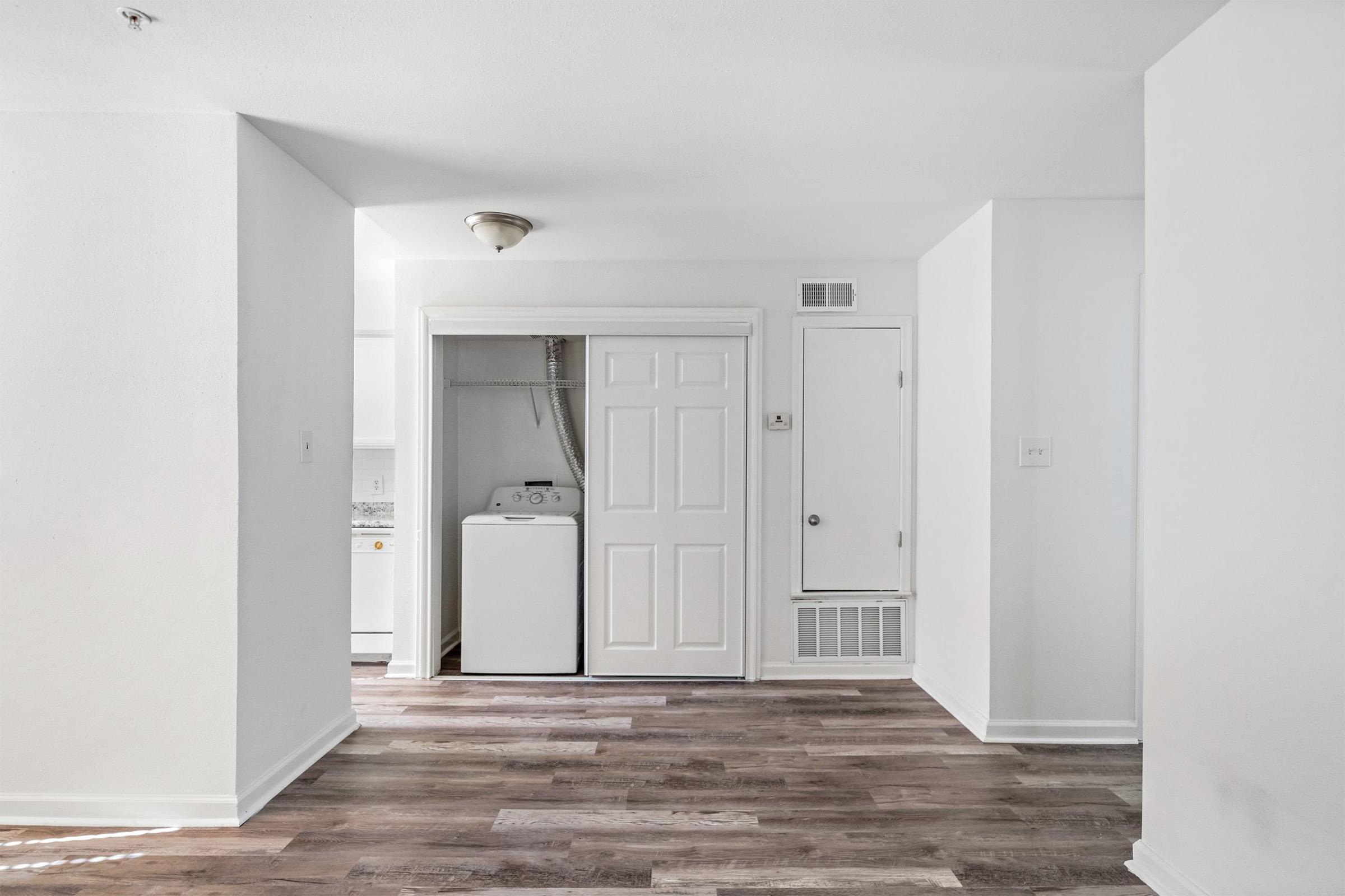 A bright, modern interior of an apartment featuring a spacious living area with light-colored walls and wood-style flooring. On the left, there's a doorway leading to a laundry area with appliances visible. The overall space is clean and well-lit, creating an inviting atmosphere.