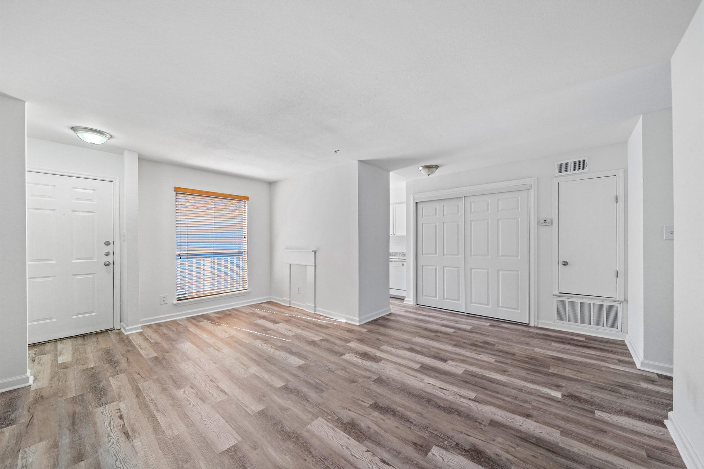 A spacious and bright interior of an apartment or room featuring light-colored walls, wood-style flooring, and a large window with blinds. The area includes a front door with a window beside it, a hallway leading to a closet, and an open layout that transitions to a kitchen area in the background.