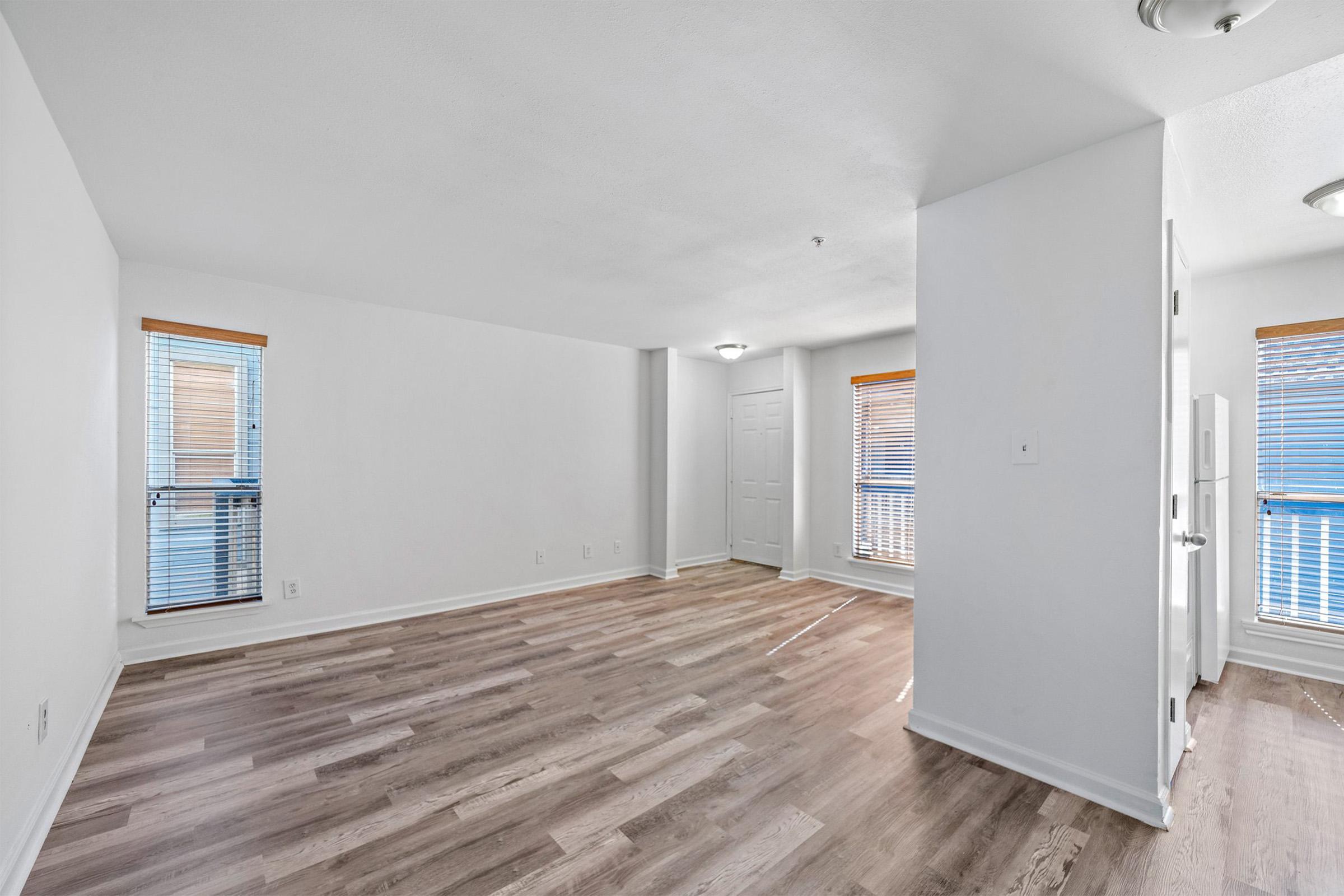 A spacious, well-lit room with light-colored walls and wooden flooring. There are two windows with blinds letting in natural light, and a door visible in the background. The overall atmosphere is clean and modern, suitable for a living area or office space.
