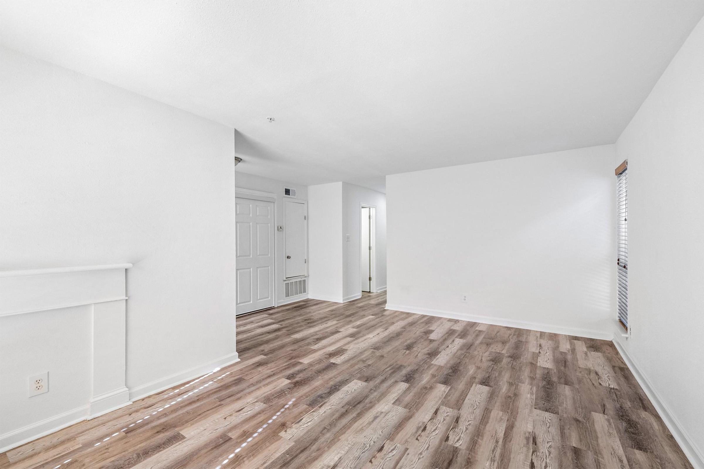 Empty living room with light-colored walls and wooden laminate flooring. A white fireplace is visible on the left, with a door leading outside to the entrance area. Natural light comes through a window on the right, highlighting the spacious, bright atmosphere of the room.