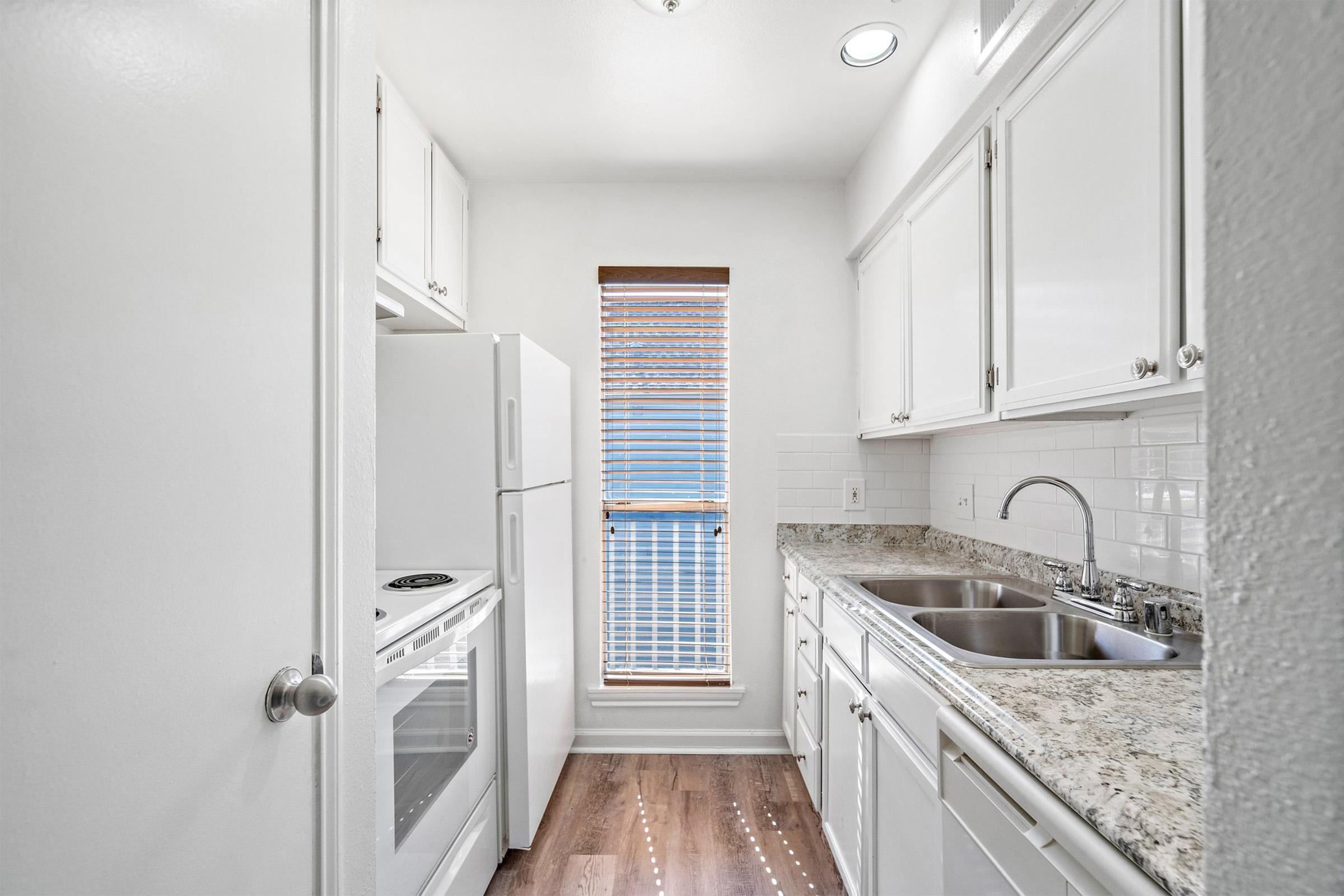 A bright, modern kitchen featuring white cabinetry, a granite countertop, stainless steel sink, and appliances including a refrigerator and oven. Natural light streams in through a window with wooden blinds, highlighting the clean, organized space and warm wood flooring.
