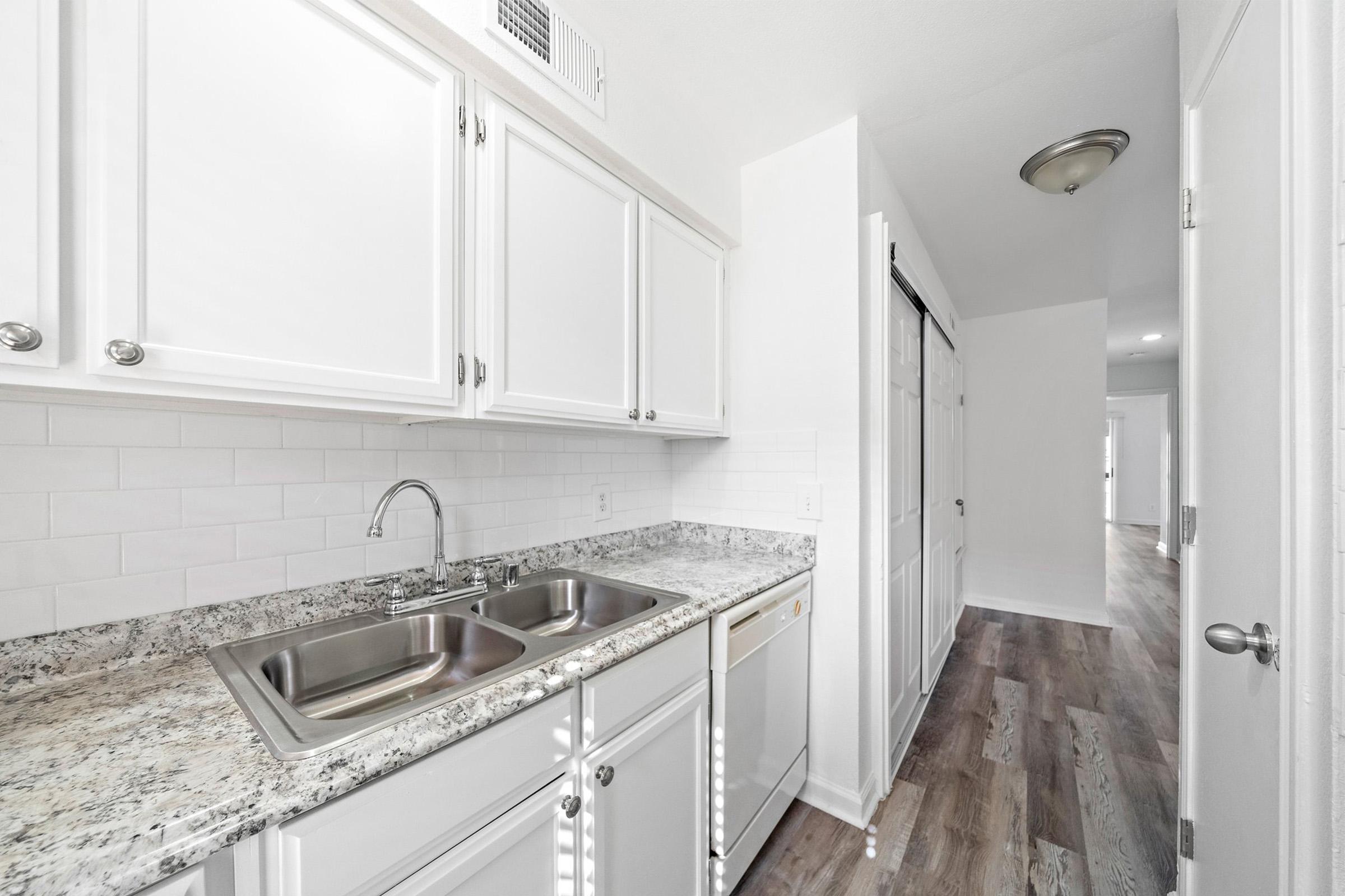 A modern, well-lit kitchen with white cabinets, a double sink, and granite countertops. The kitchen features an adjacent hallway with light-colored wood flooring and a doorway leading to another room. The walls are painted white, creating a clean and spacious atmosphere.