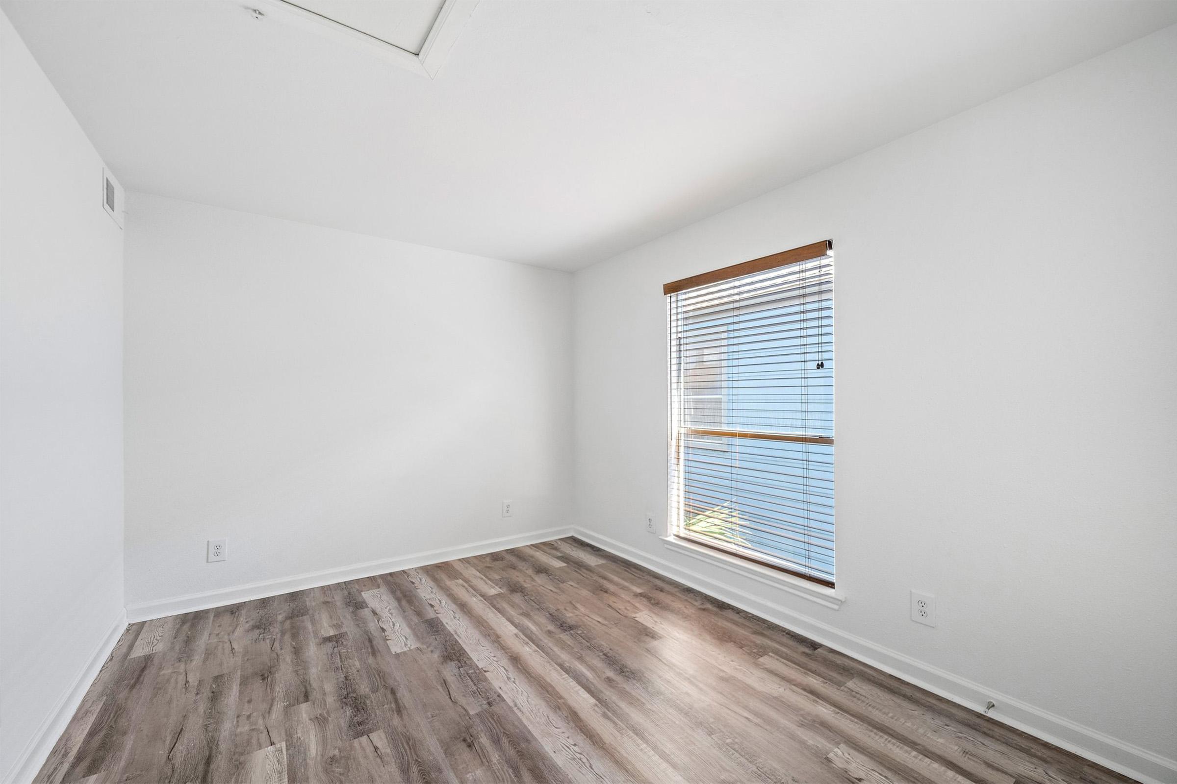 Empty room with white walls and a window covered by blinds, allowing natural light to enter. The floor is made of light-colored wood, and there are no furnishings or decorations in the space, creating a minimalist and open atmosphere.