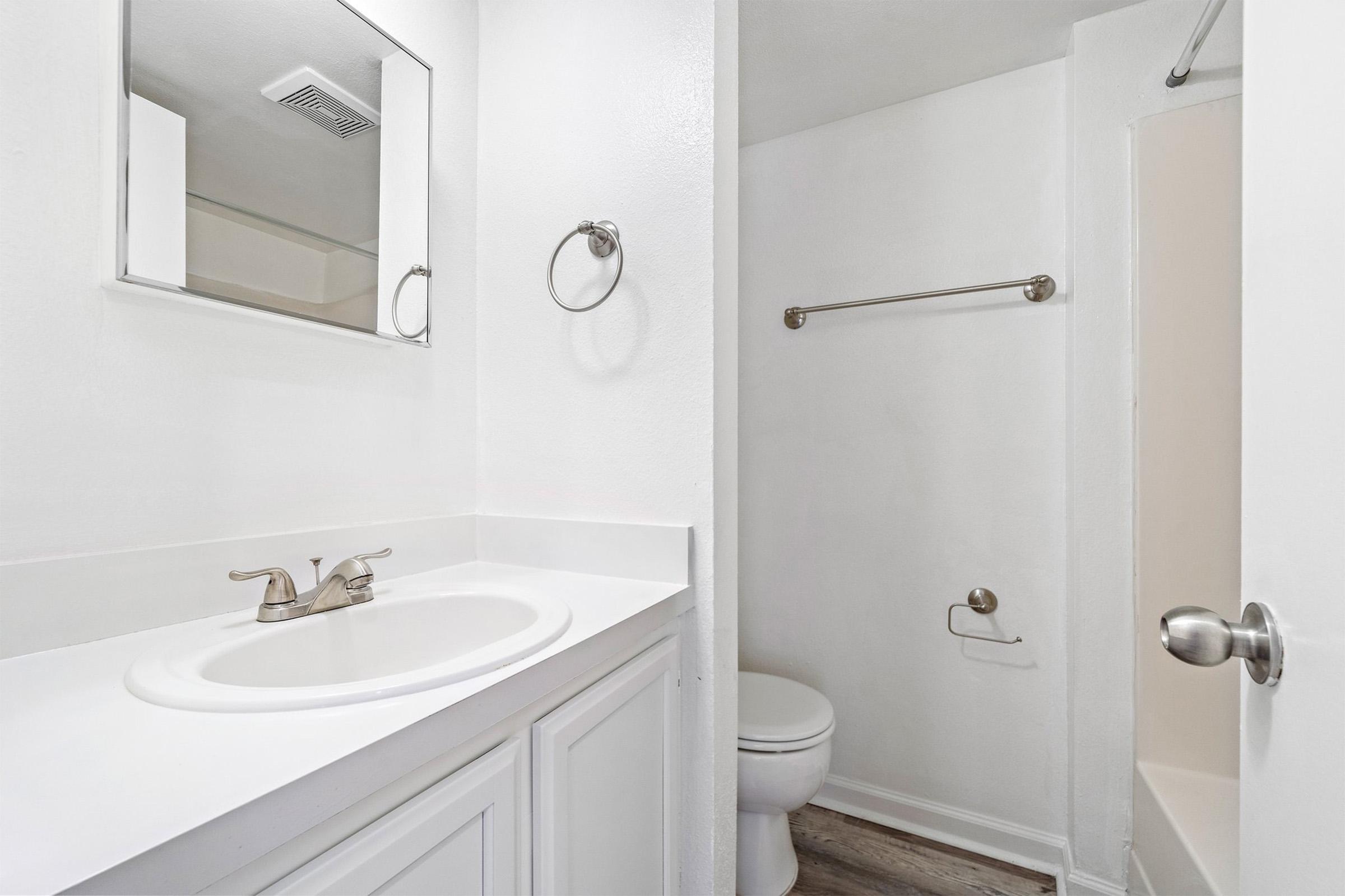 A clean, modern bathroom featuring a white sink with a silver faucet, a large mirror above, and a toilet. The walls are painted white, and there is a towel ring on the wall. A shower stall is visible on the right, with a neutral-colored floor. The overall design is bright and minimalist.